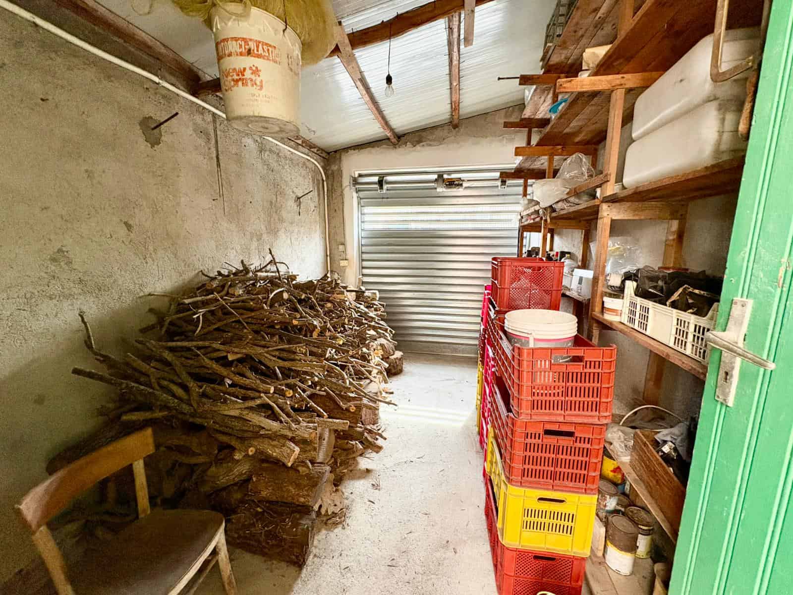 Palo di legna e attrezzi da giardinaggio in un garage o deposito, con scaffalature in legno e porta in metallo, ambiente ordinato e funzionale per il fai-da-te e il giardinaggio.