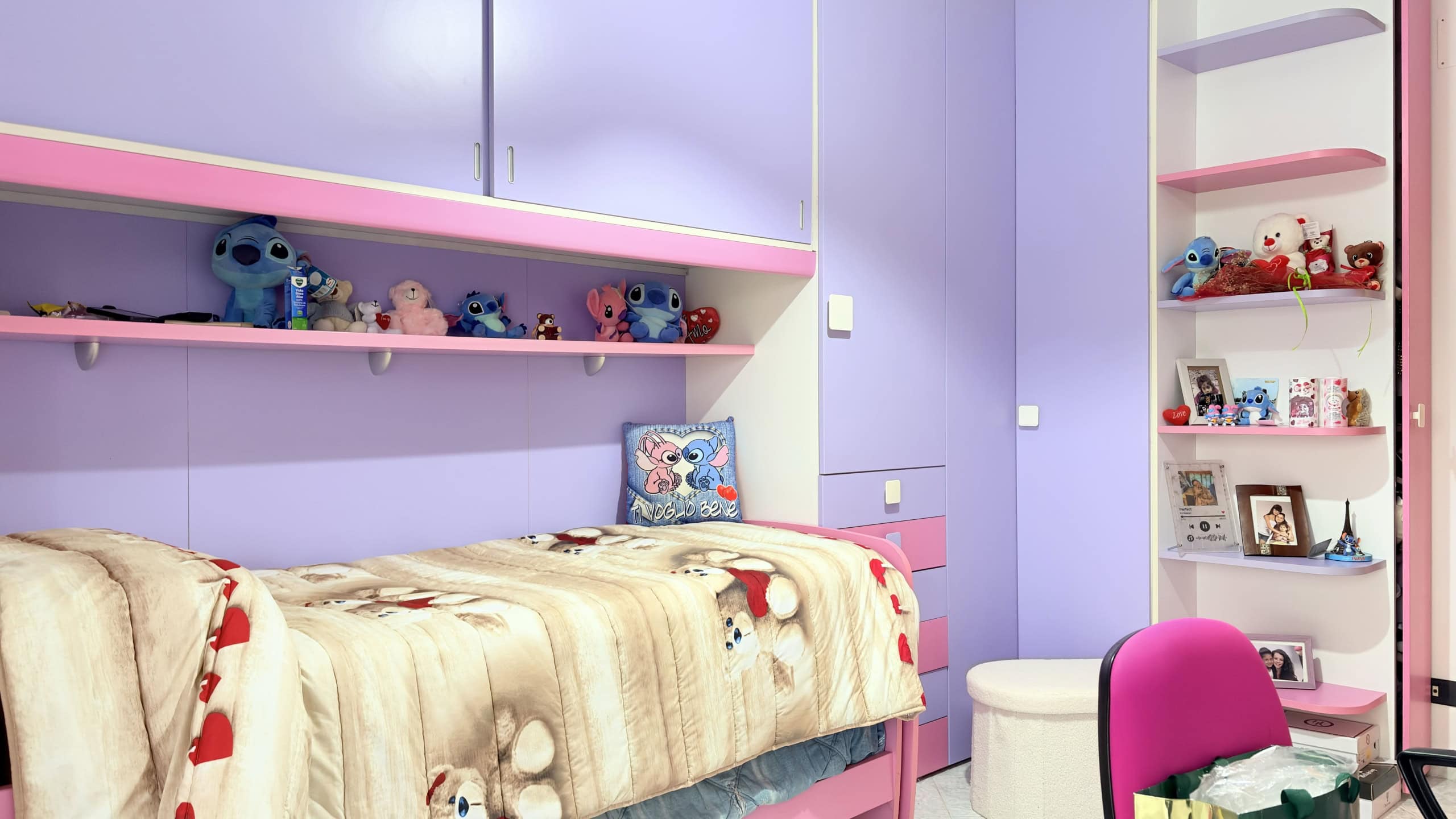 Letto singolo con copriletto decorato, in una stanza color lavanda con scaffali bianchi pieni di giocattoli e foto, ambiente accogliente e luminoso, ideale per bambini o adolescenti.