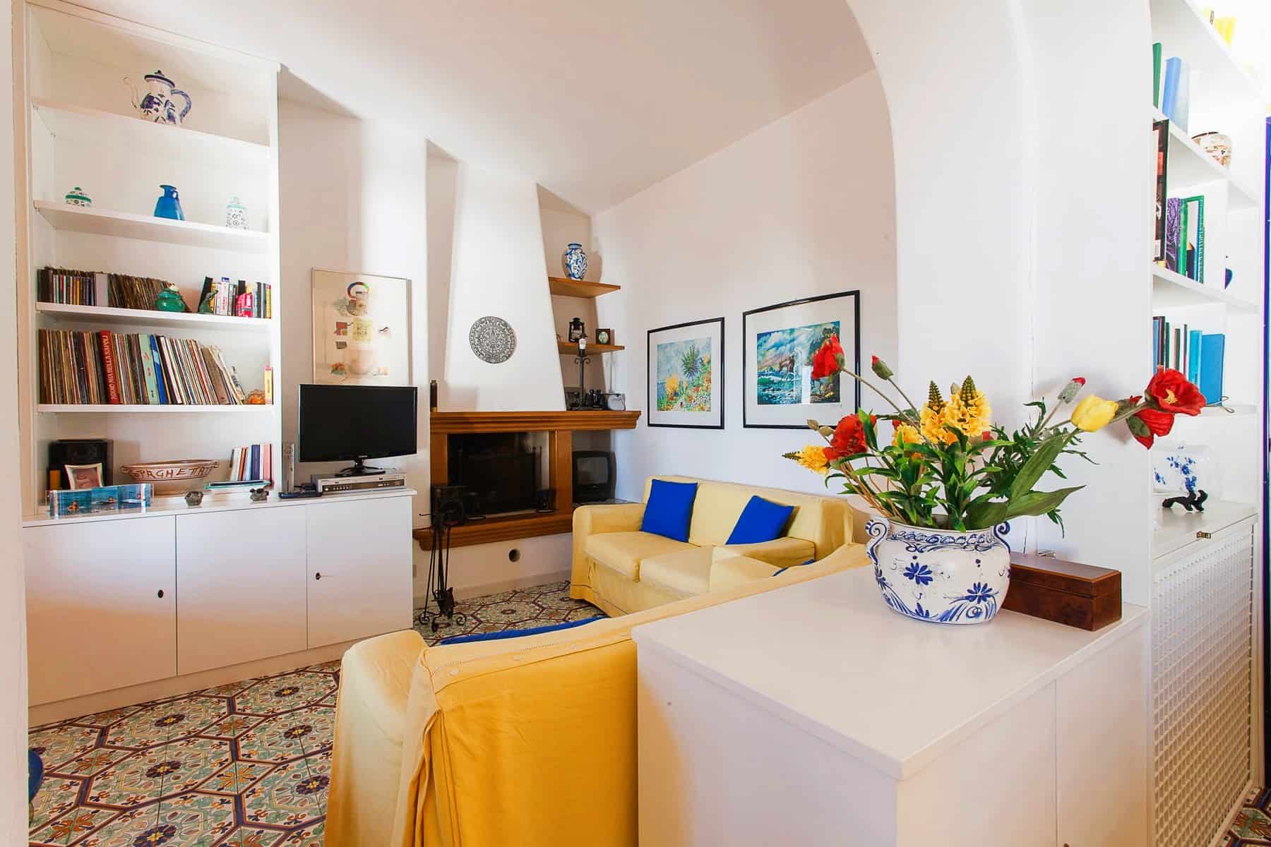 Gli immobili di NextCasa: soggiorno luminoso con pareti bianche, librerie piene di libri, divani gialli e decorazioni colorate, ideale per vivere in ambienti accoglienti e funzionali.