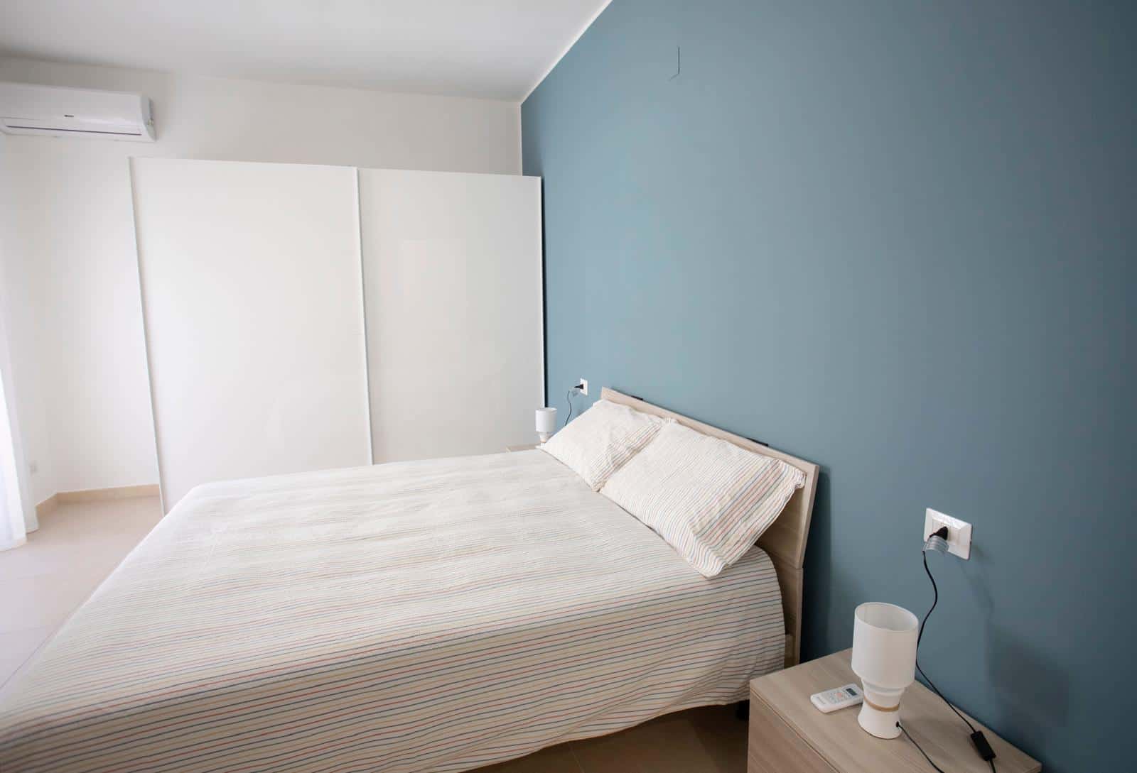 L'immagine mostra una camera da letto moderna con pareti bianche e una parete blu, letto matrimoniale con lenzuola a righe e due cuscini, comodino con lampada e diffusore, ambiente luminoso e accoglie.