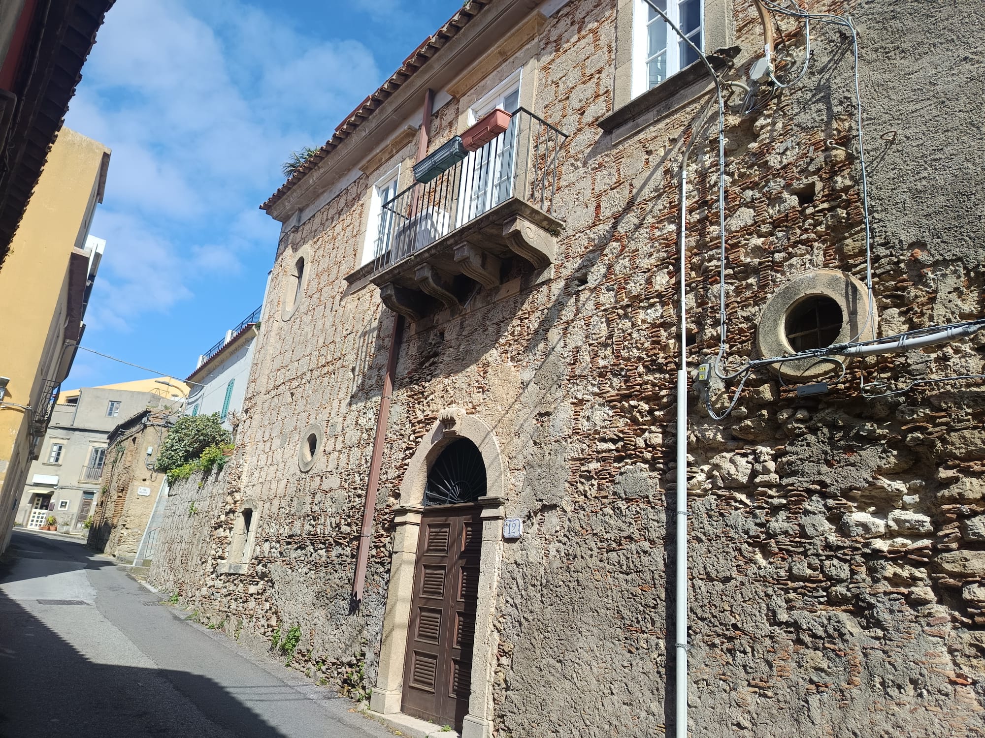 Vecchia casa in pietra con balcone in ferro battuto e porta in legno, situata in una strada stretta e pittoresca di un quartiere storico italiano.