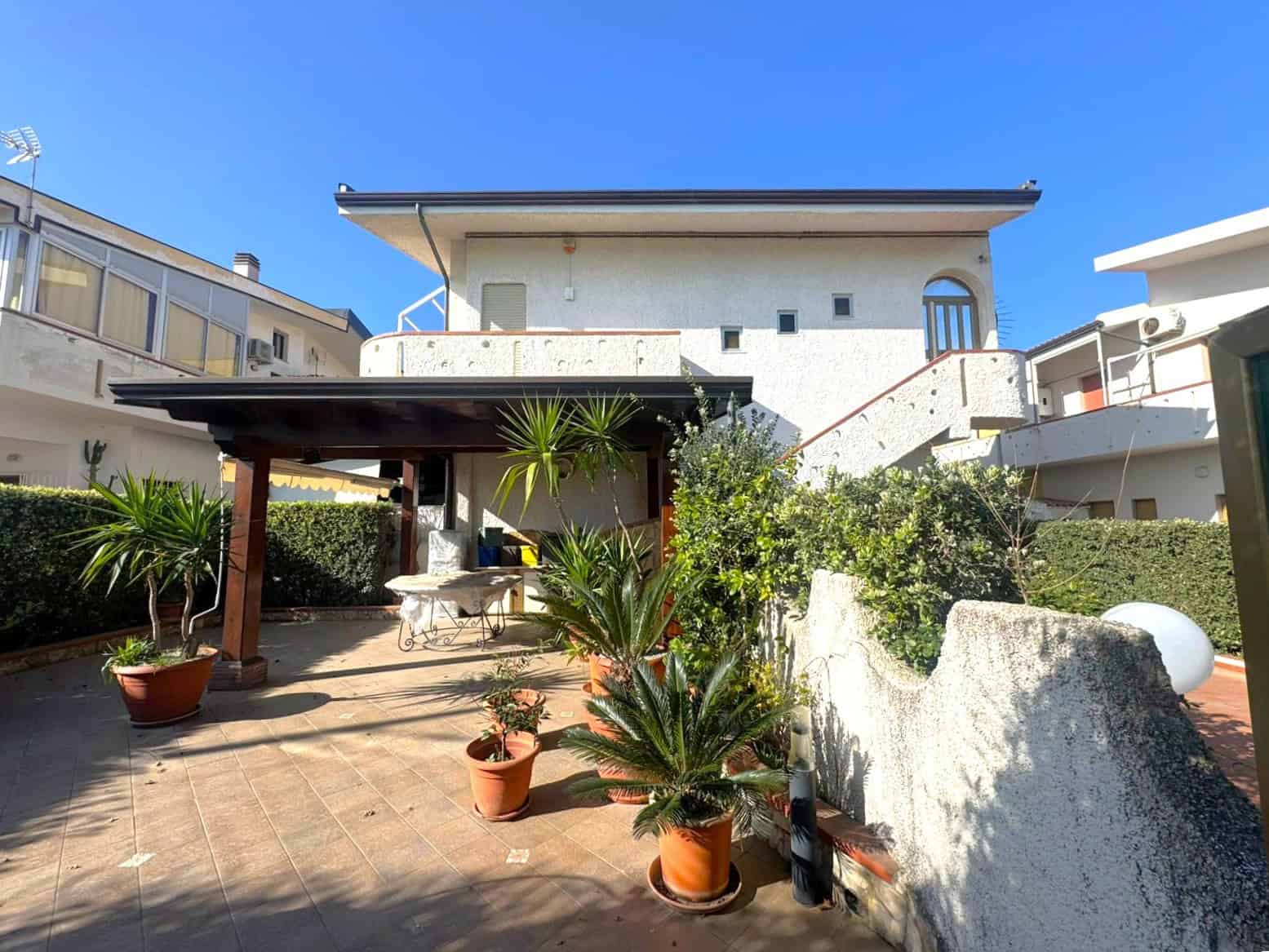Esterno di una casa con patio e piante in vaso, sotto un cielo azzurro, in zona residenziale, ideale per relax e convivialità.