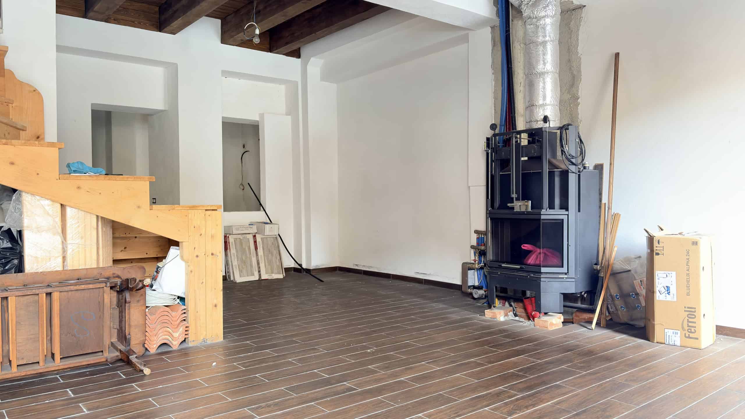 Ampia stanza abitativa in fase di ristrutturazione con pavimento in gres effetto legno, camino moderno e pareti bianche luminose, pronta per essere personalizzata.