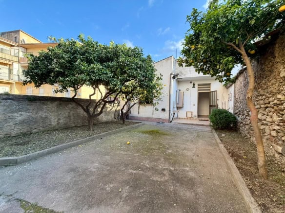 Piante e cortile esterno di una casa in stile mediterraneo con pareti bianche e un'area di parcheggio in pietra, sotto un cielo azzurro con nuvole sparse, ambiente tranquillo e accogliente.