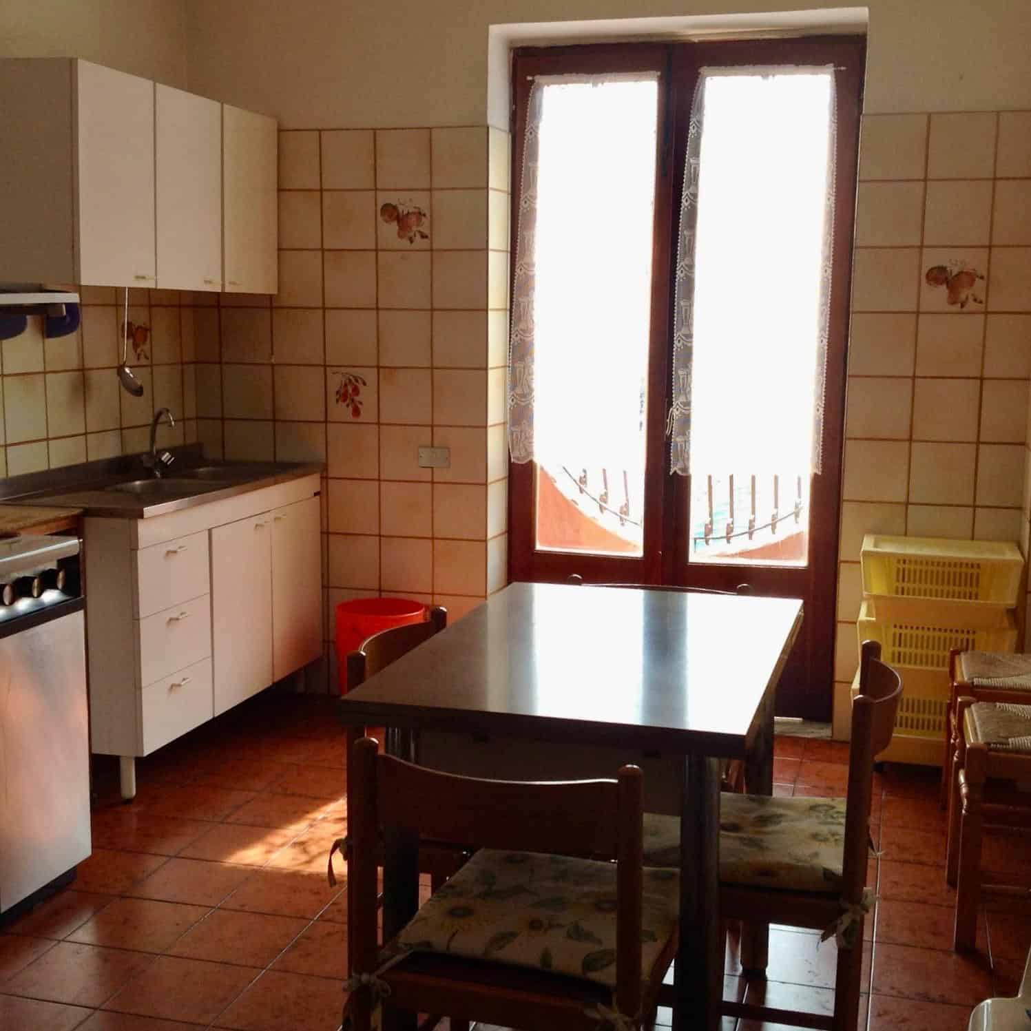Cucina semplice con tavolo e sedie vicino a porta finestra, illuminata dalla luce naturale.