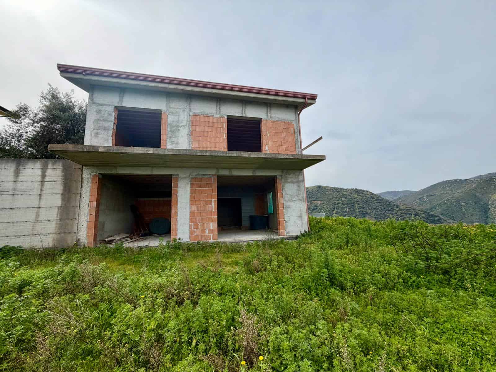 Casa in costruzione con struttura in cemento e mattoni, situata in un'area verde con vista sulle colline, pronta per essere completata e personalizzata.
