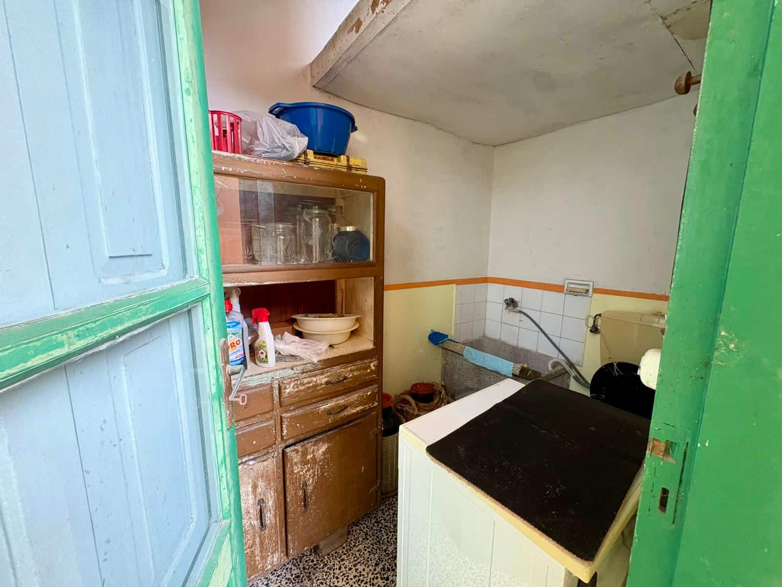 Lavatrice e scaffale in una stanza di servizio con pareti bianche e soffitto inclinato, arredi usurati e oggetti di uso quotidiano, ambiente funzionale e pratico per le faccende domestiche.