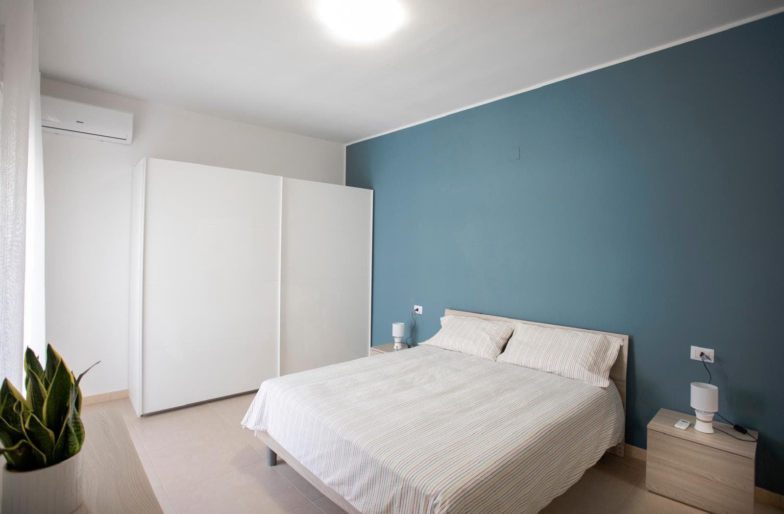 Camera da letto moderna con parete blu, letto matrimoniale, armadio bianco e arredi minimalisti, ambiente luminoso e accogliente, ideale per soggiorni confortevoli.