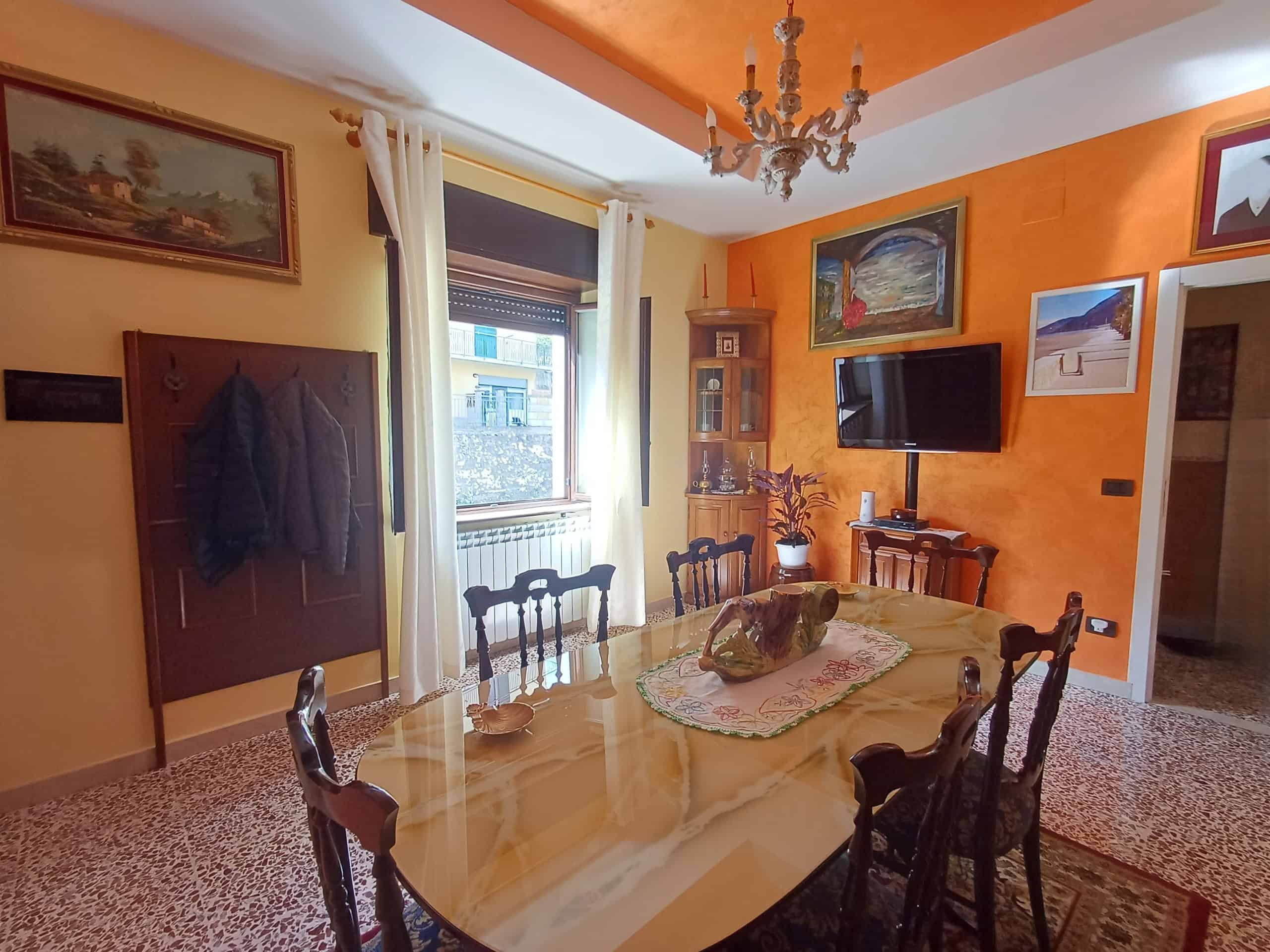 Pannello di ingresso con appendiabiti e quadri alle pareti, soggiorno accogliente con pareti arancioni, tavolo in legno con sedie, televisore a schermo piatto e decorazioni floreali, ambiente luminoso.