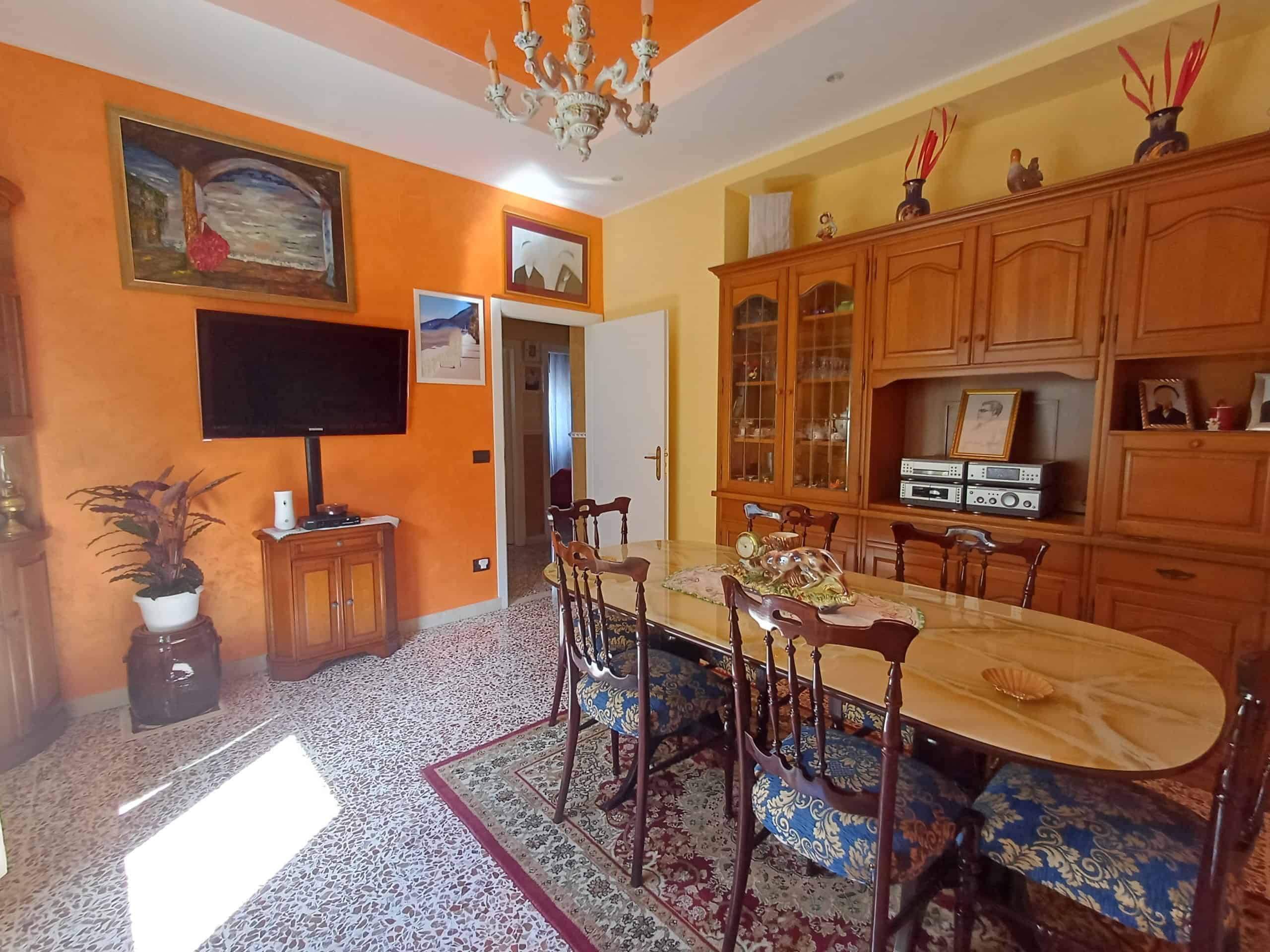 Una sala da pranzo accogliente con pareti arancioni, un grande tavolo in legno con sedie imbottite, un mobile in legno con vetrine e un televisore a schermo piatto, decorata con quadri e un lampadario.