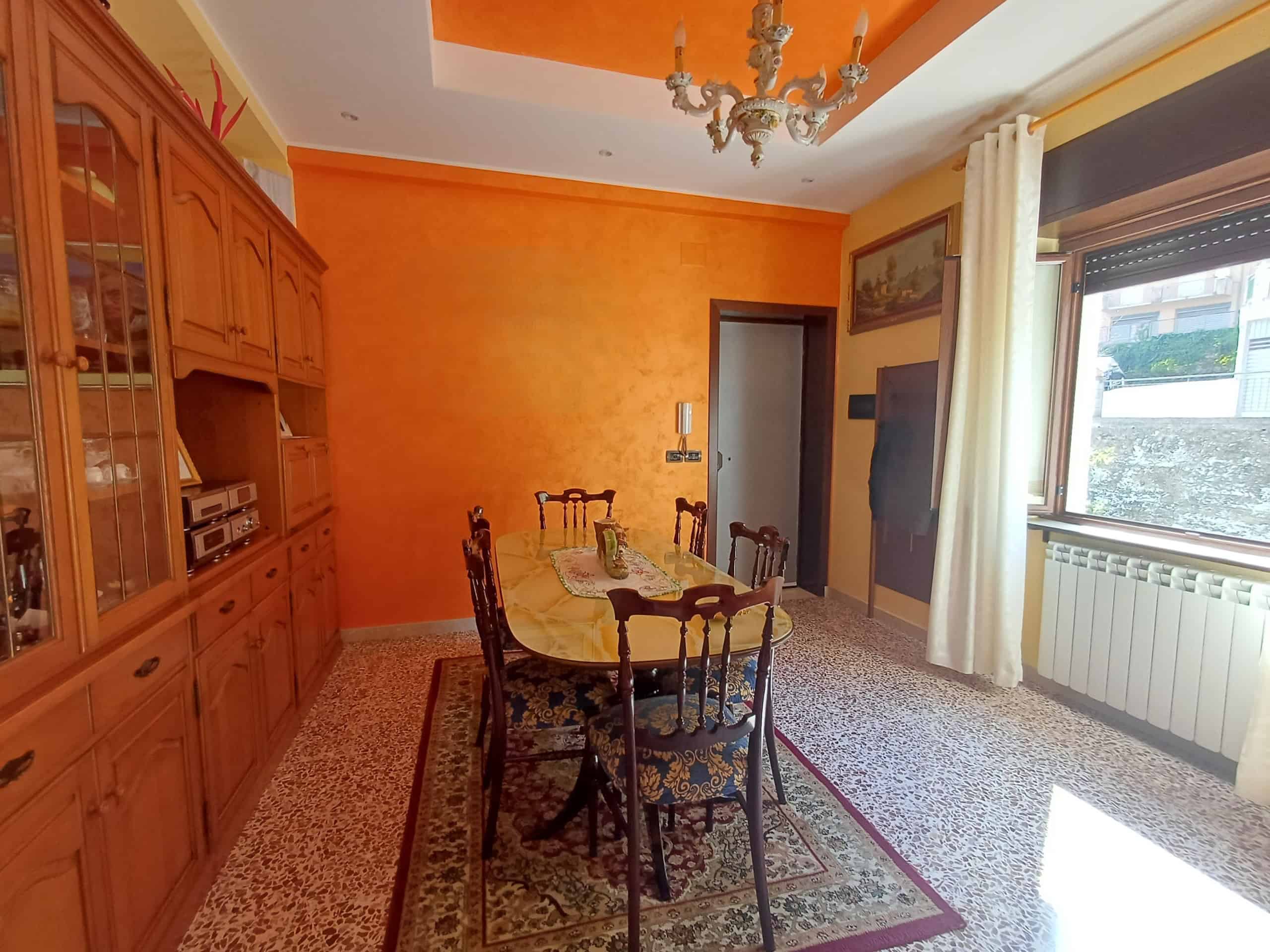 Una sala da pranzo accogliente con pareti arancioni, mobili in legno, un tavolo con sedie, un lampadario elegante e una grande finestra con tende bianche, ideale per vivere momenti conviviali.
