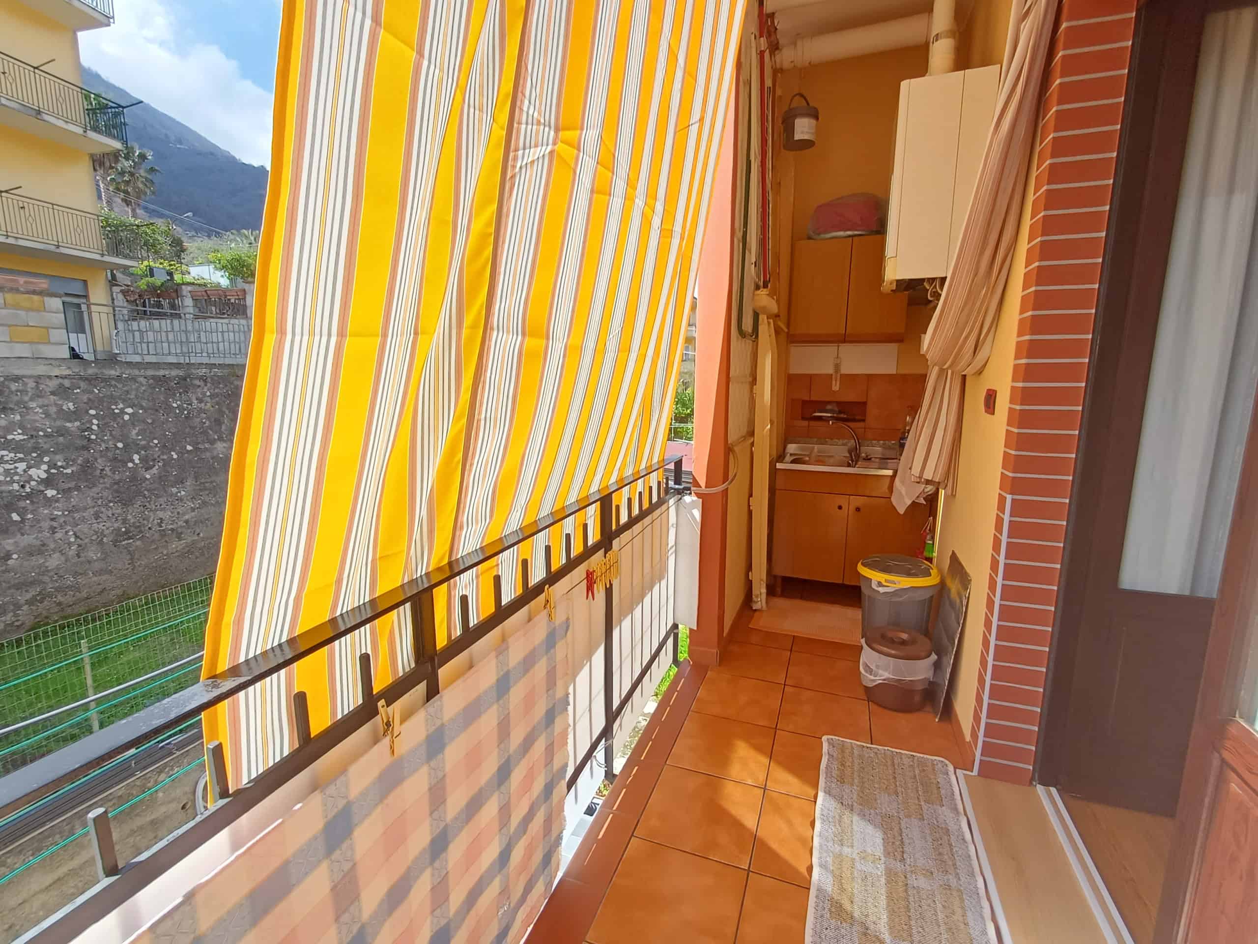 Balcone di un appartamento con tende a righe gialle e bianche, vista su un paesaggio montano, con arredi esterni e accessori per il relax e la praticità quotidiana.
