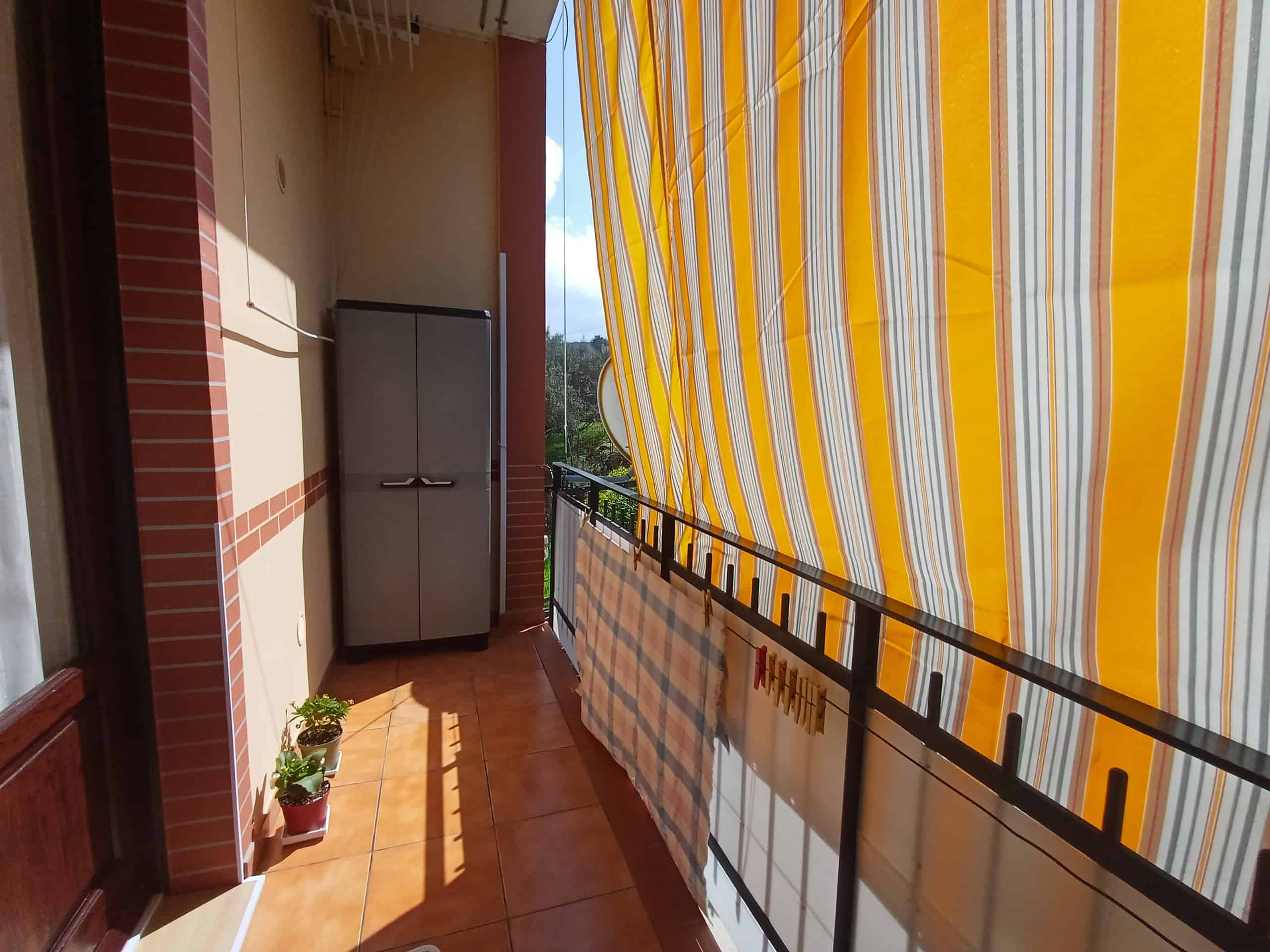 Balcone di un appartamento con tenda parasole gialla e bianca, frigorifero e piante in vaso, vista panoramica sul cielo e sulla natura circostante, ambiente luminoso e accogliente.