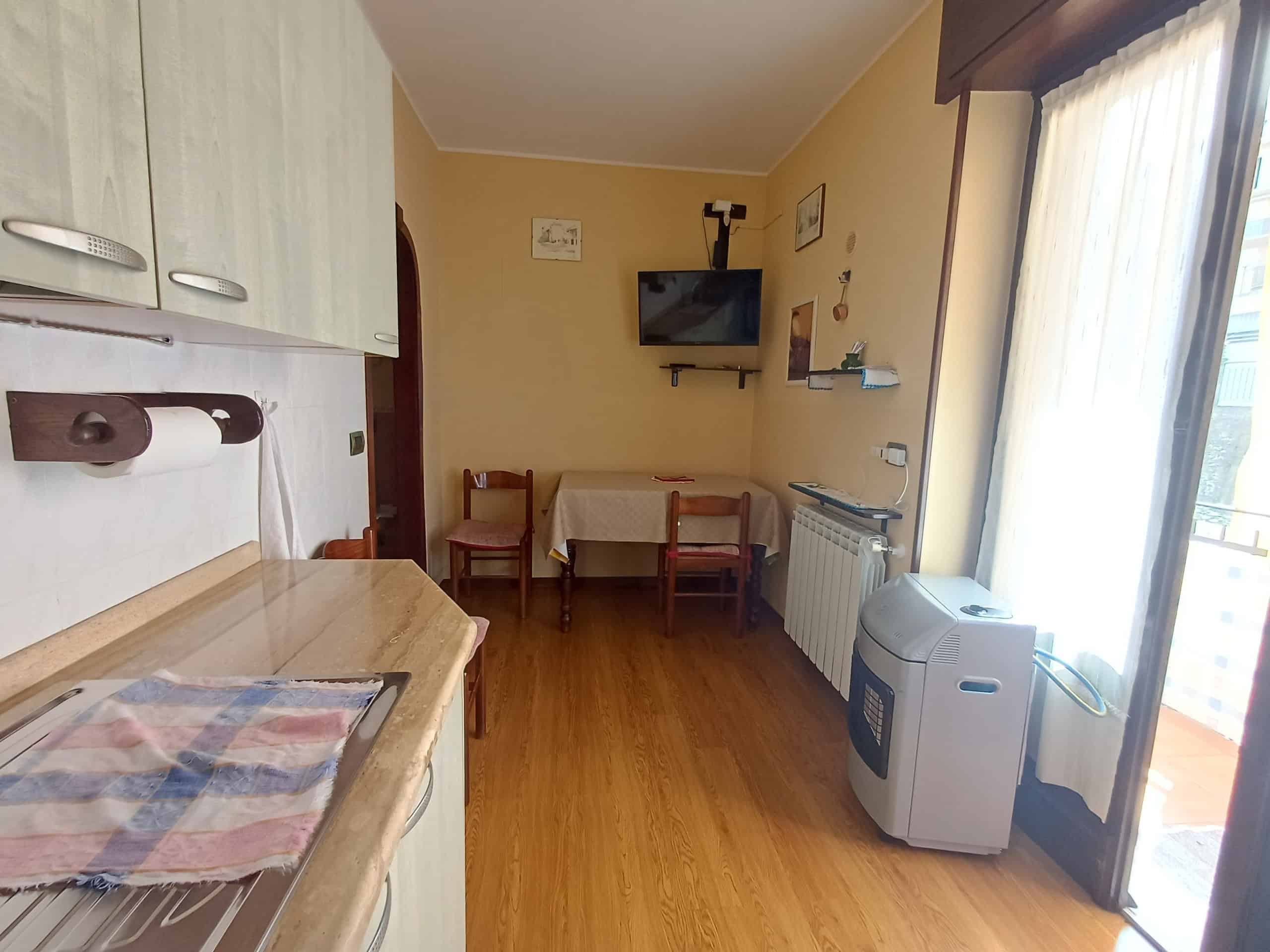 Ampio soggiorno con balcone, arredato con tavolo, sedie, cucina compatta e TV, ideale per relax e convivialità, con accesso diretto alla terrazza soleggiata.