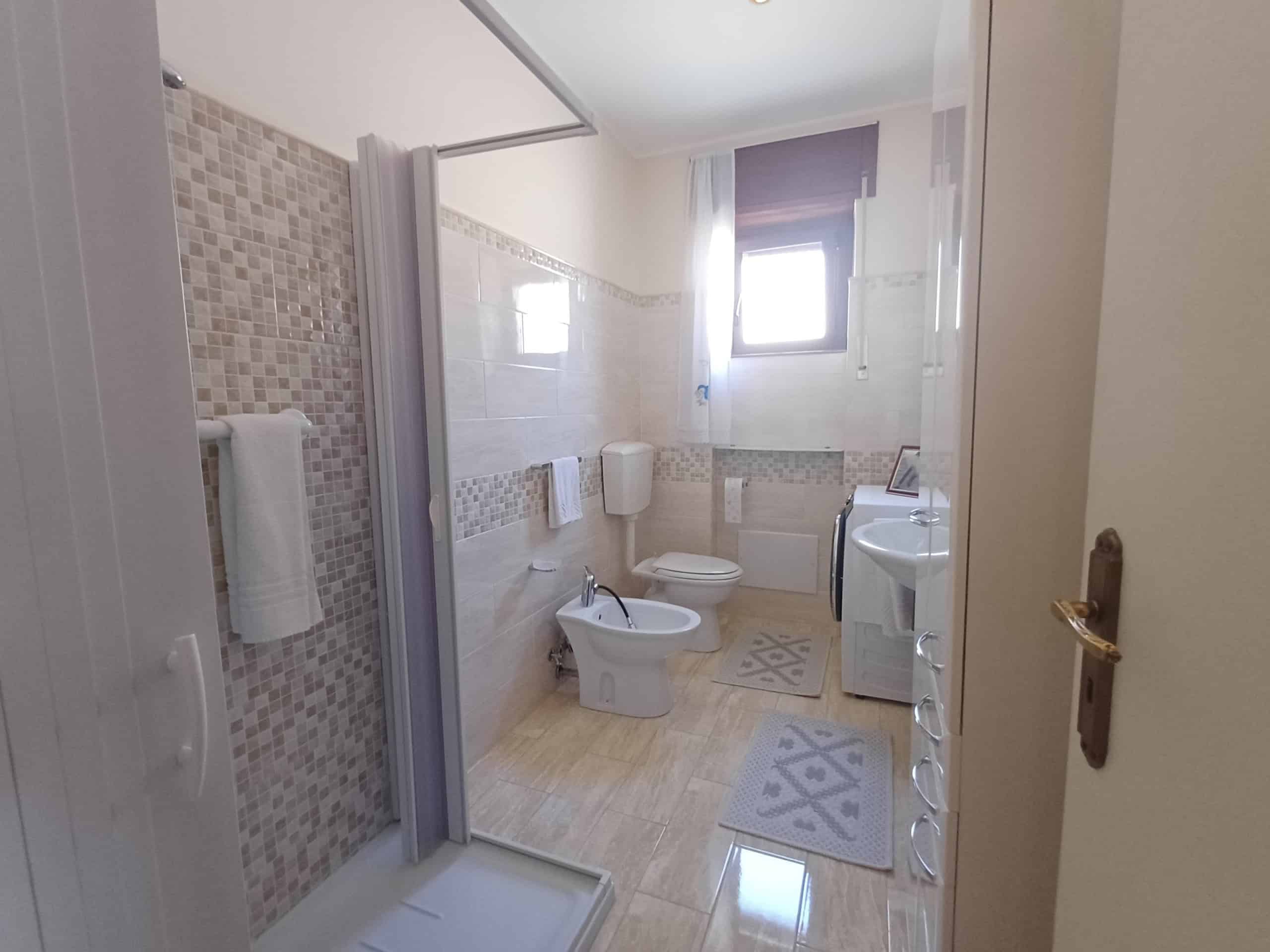 Il bagno moderno con doccia, bidet e WC, illuminato da una finestra, con arredi funzionali e pavimento in piastrelle beige, ideale per case in vendita o affitto a NextCasa.