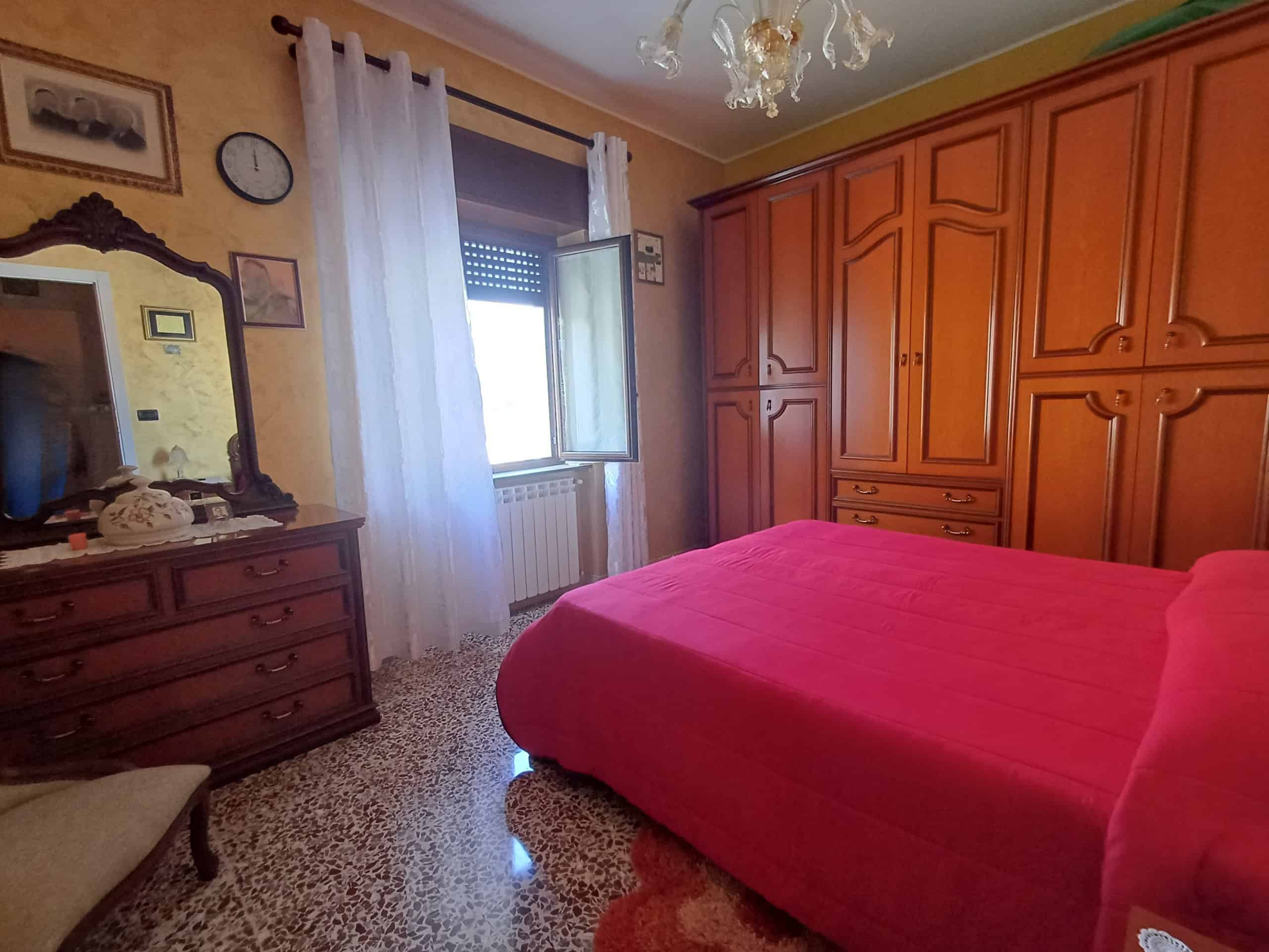 Ampio camera da letto con armadio a muro in legno, letto matrimoniale con copriletto rosso, finestra con tende bianche, arredi classici, pareti colorate, lampadario e specchio decorativo.