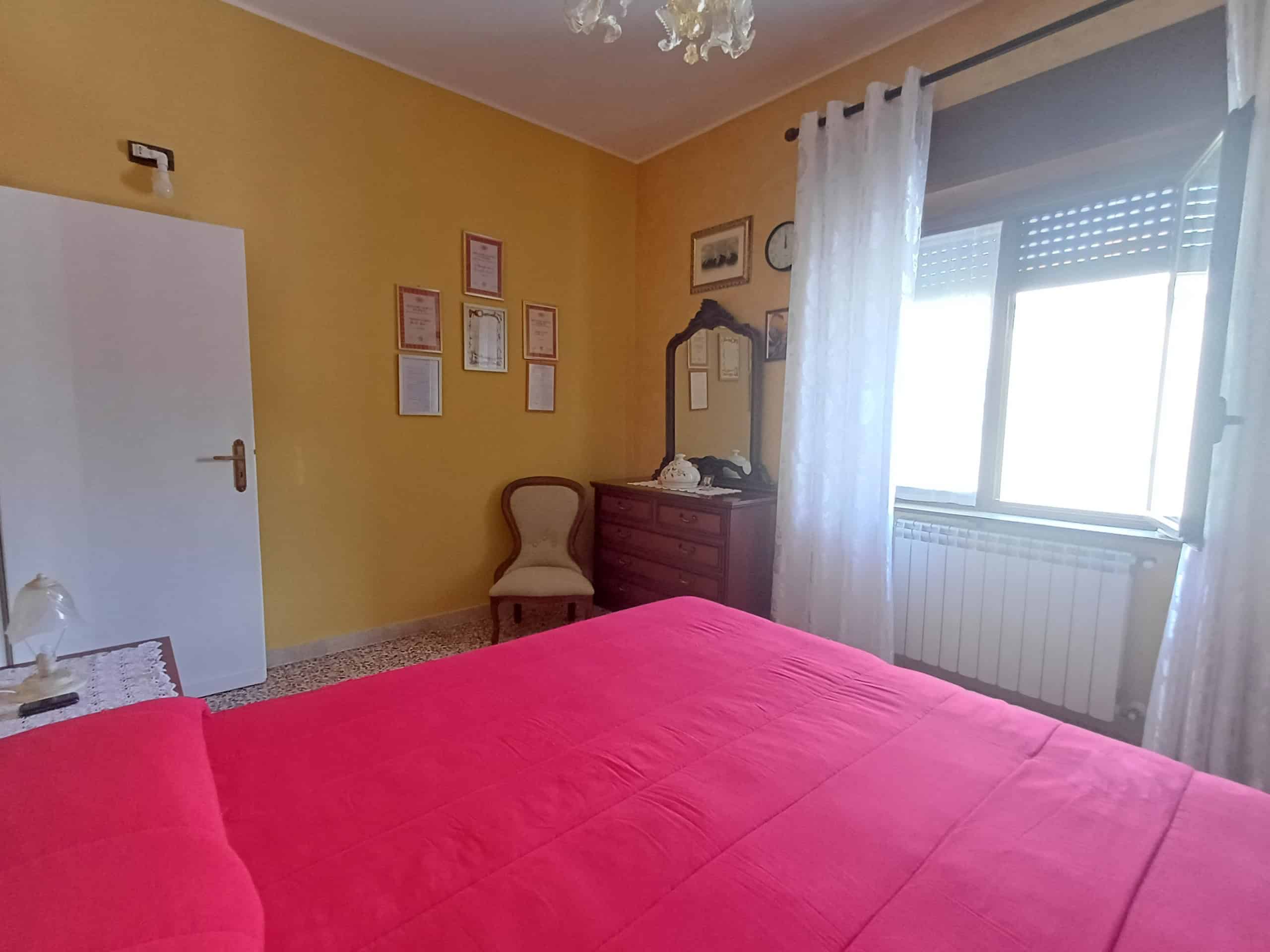 Letto matrimoniale con lenzuola rosa in camera da letto luminosa, decorata con quadri e specchio, finestra con tende bianche, ambiente accogliente e ben arredato, ideale per soggiorni confortevoli.