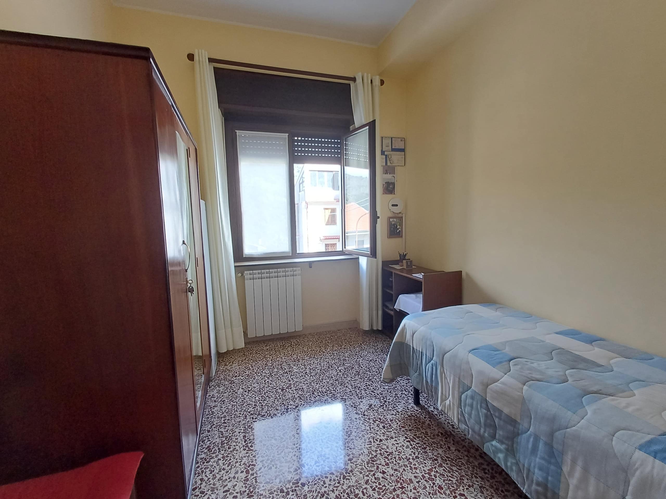 Letto singolo in camera con finestra luminosa, arredamento semplice e pavimento in piastrelle. Ideale per affitti brevi o studenti, con spazio funzionale e atmosfera accogliente.