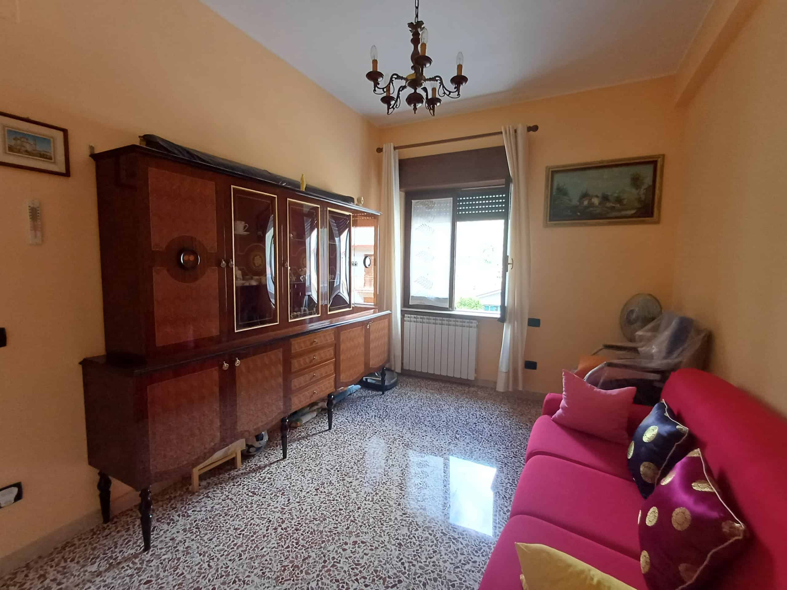 Un mobile antico in legno in soggiorno con pareti color crema, finestra con tende bianche e divano rosso con cuscini decorativi, ambiente accogliente e luminoso.