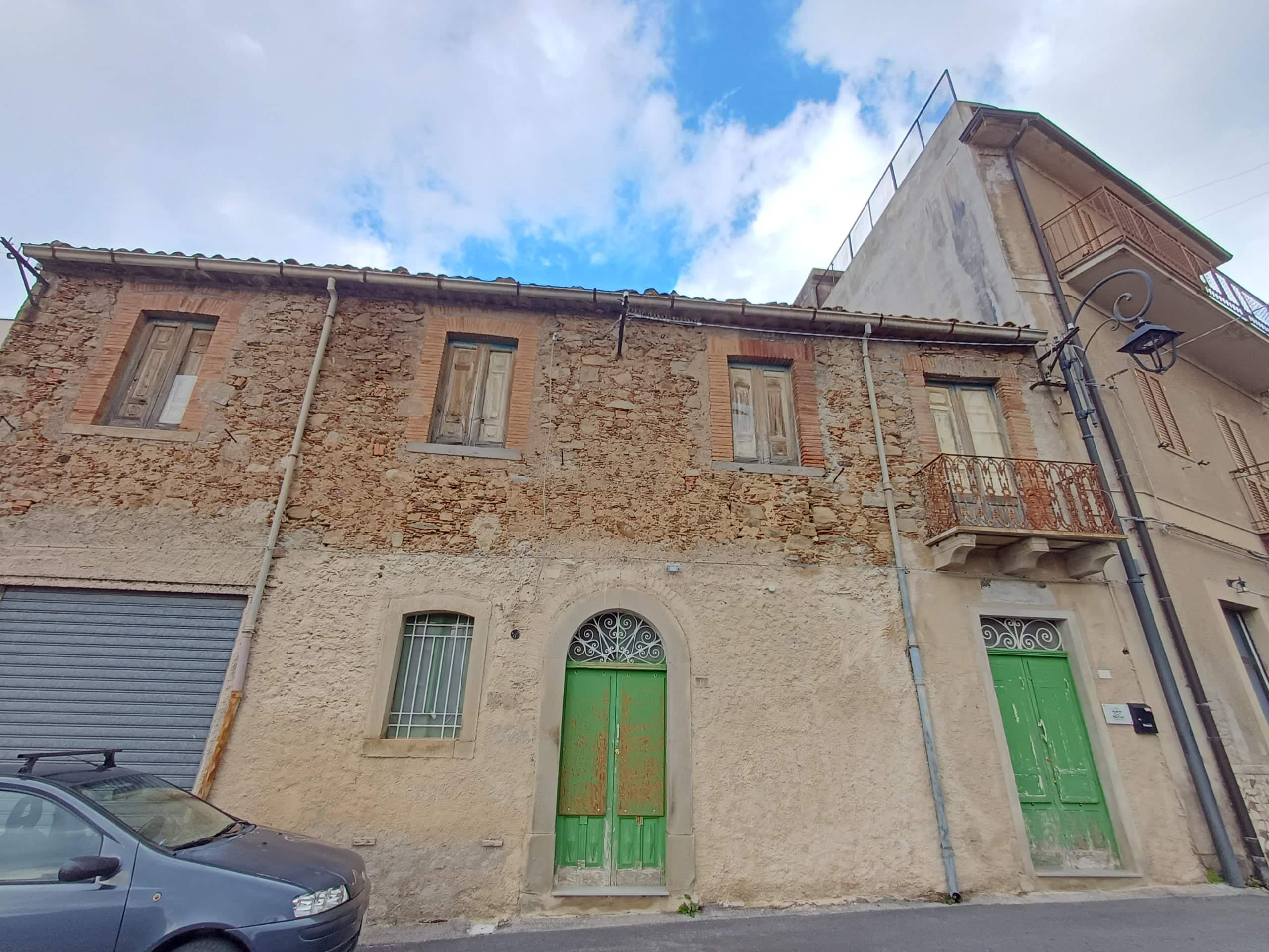 Porte in legno verde e facciata in pietra e intonaco di un edificio storico in centro città, con auto parcheggiata davanti e cielo nuvoloso.