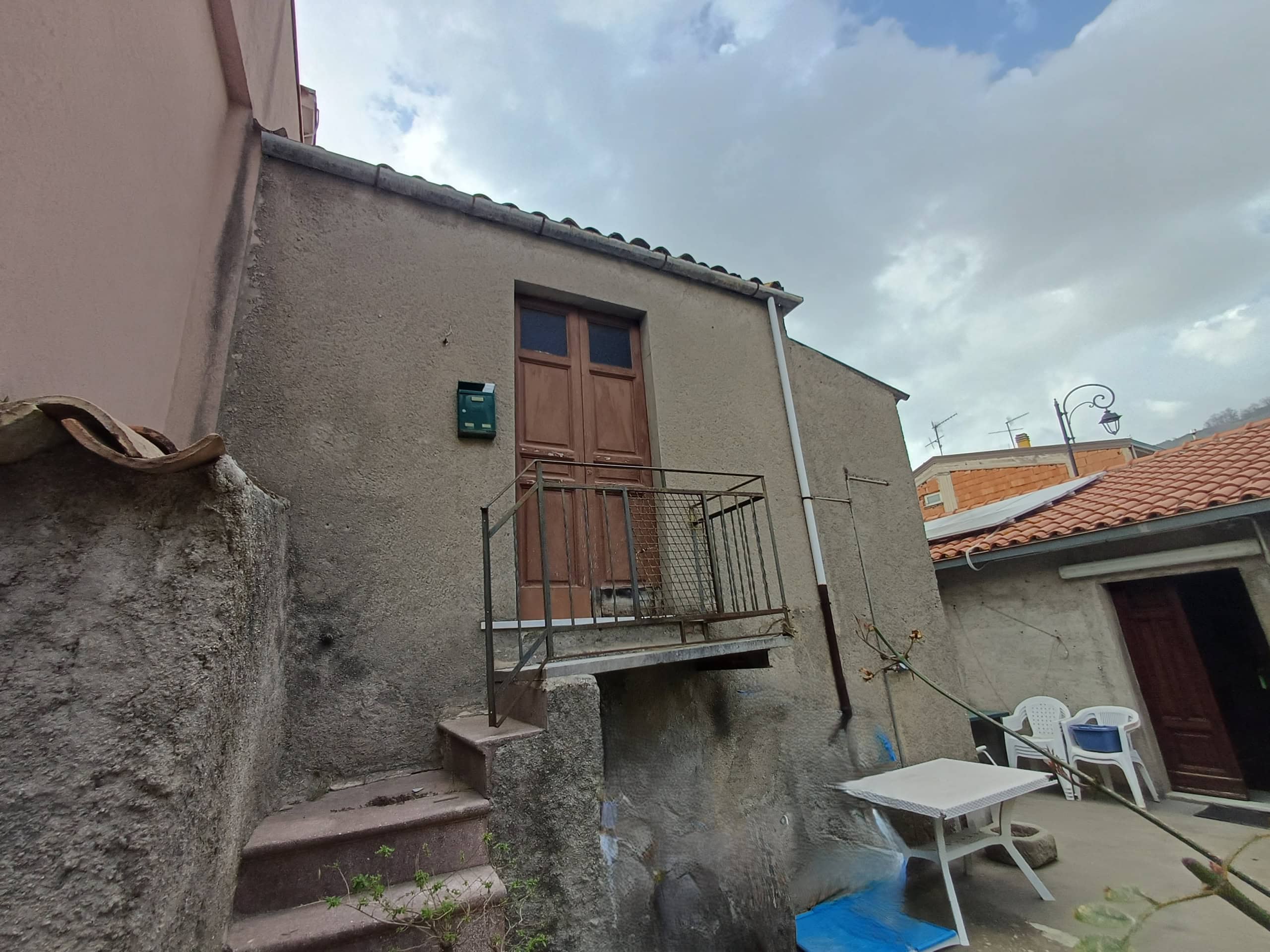 Porte in legno su un edificio con balcone in metallo, vista dall'esterno di una casa in stile rustico con pareti in pietra e cemento, cielo nuvoloso, ambiente esterno con mobili e oggetti vari.