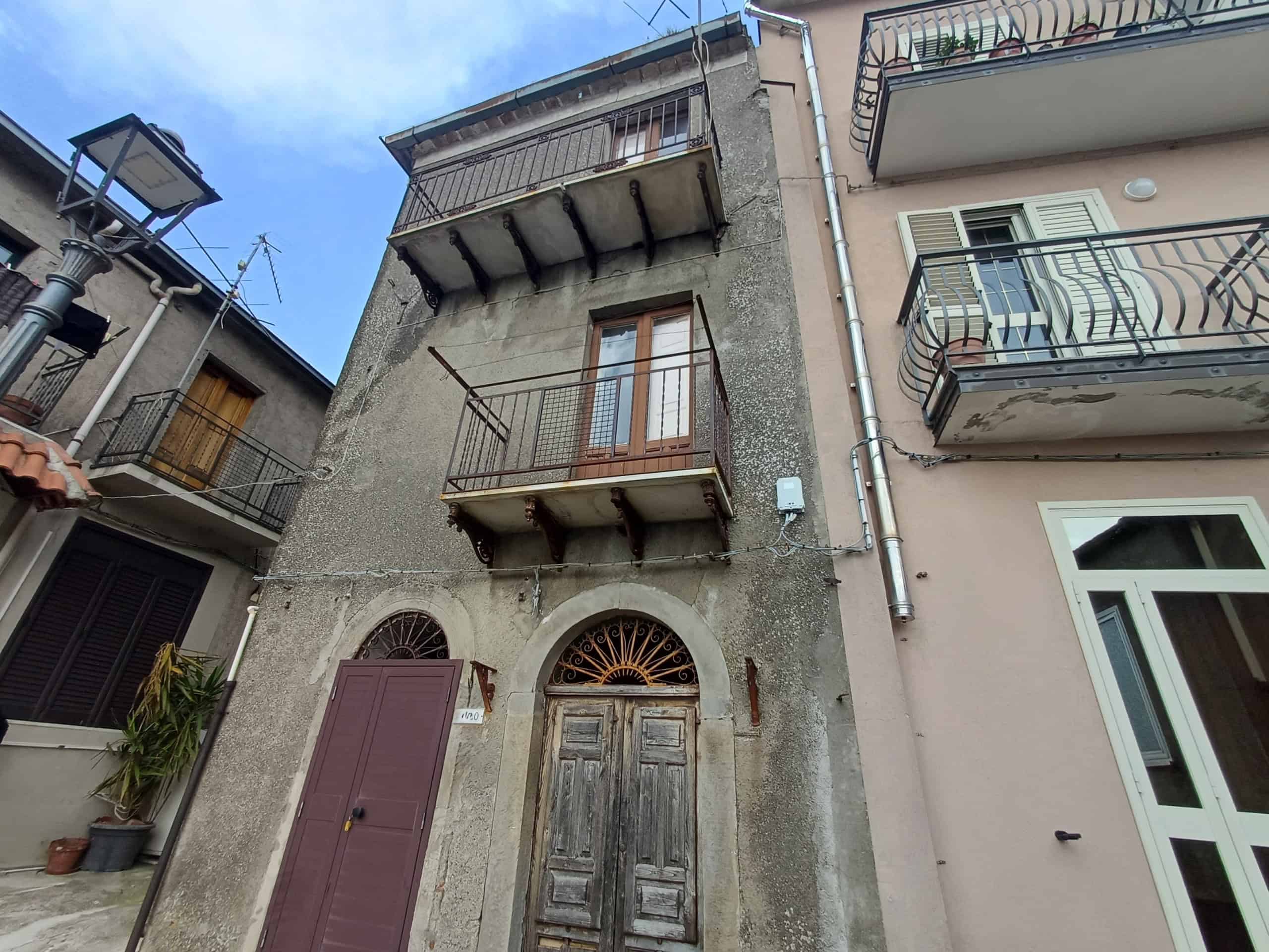 Antico edificio con balcone in ferro battuto e porta in legno, situato in zona urbana con altre abitazioni vicine, caratterizzato da un aspetto storico e autentico.