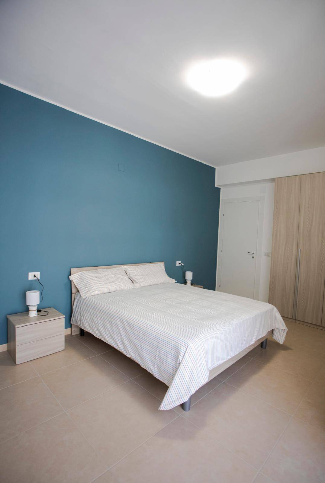 Letto matrimoniale in camera moderna con parete blu, comodini e lampade, ambiente luminoso e accogliente, ideale per affitti turistici o residenziali, arredamento minimalista e funzionale.