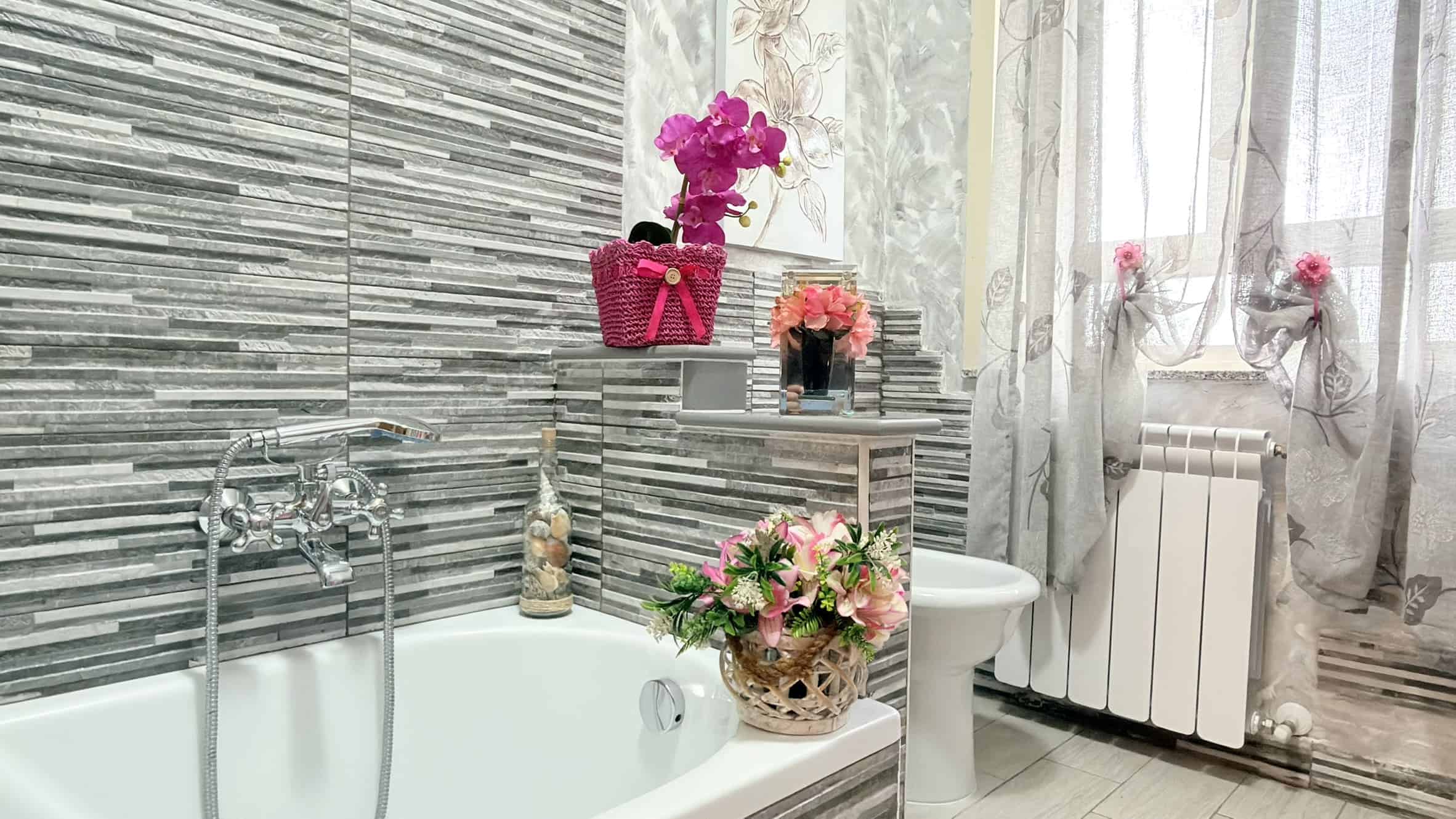 Vasca da bagno moderna con pareti in piastrelle grigie e decorazioni floreali, ambiente luminoso e accogliente, ideale per il relax e il benessere quotidiano.