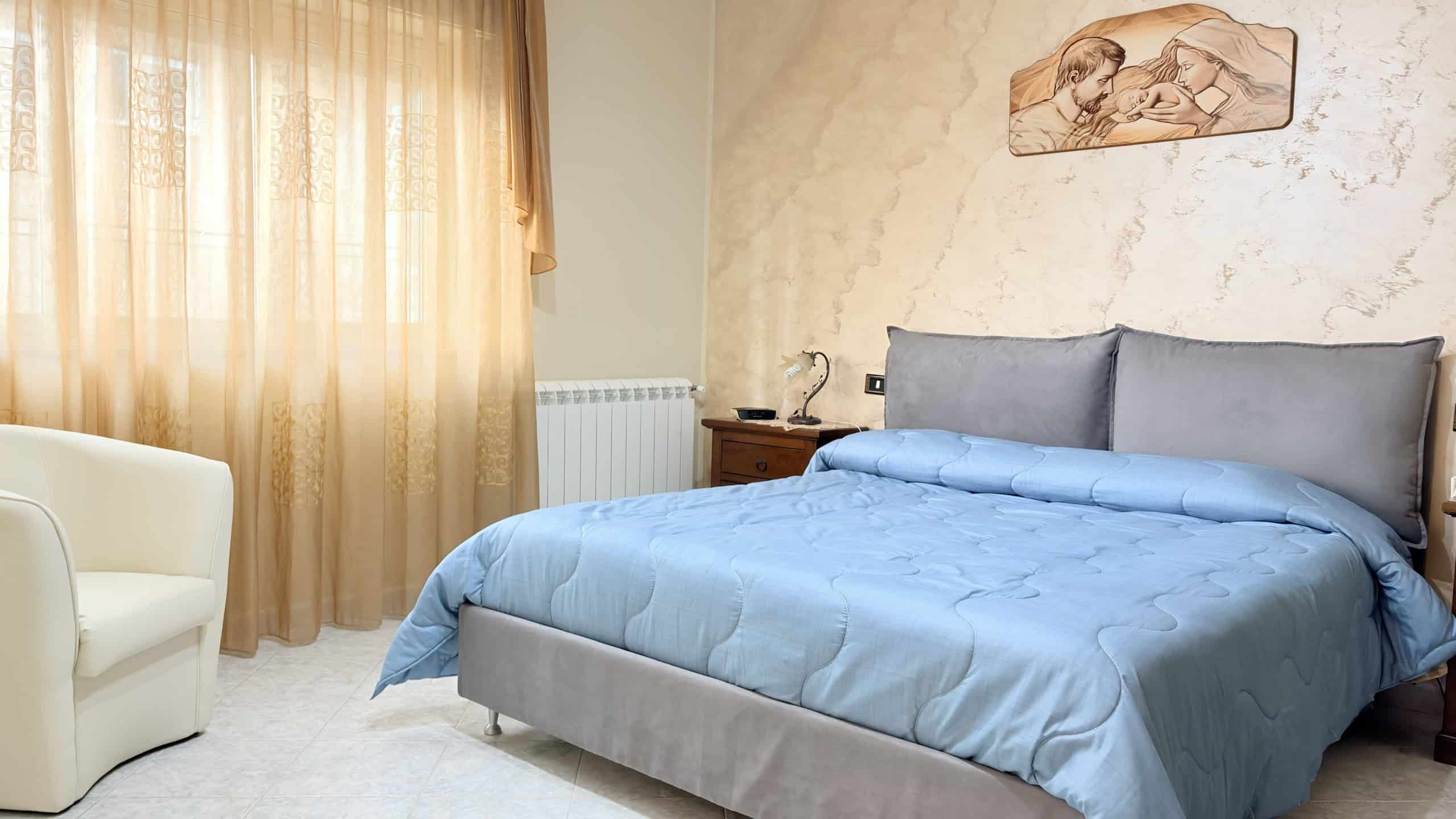 Letto matrimoniale con lenzuola azzurre in una camera da letto luminosa e accogliente, con tende beige e pareti decorate.