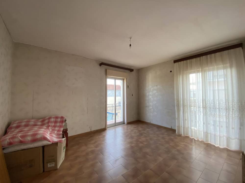 Ampia stanza luminosa con pareti neutre, pavimento in ceramica e grandi finestre con tende leggere, ideale per soggiorno o zona living.