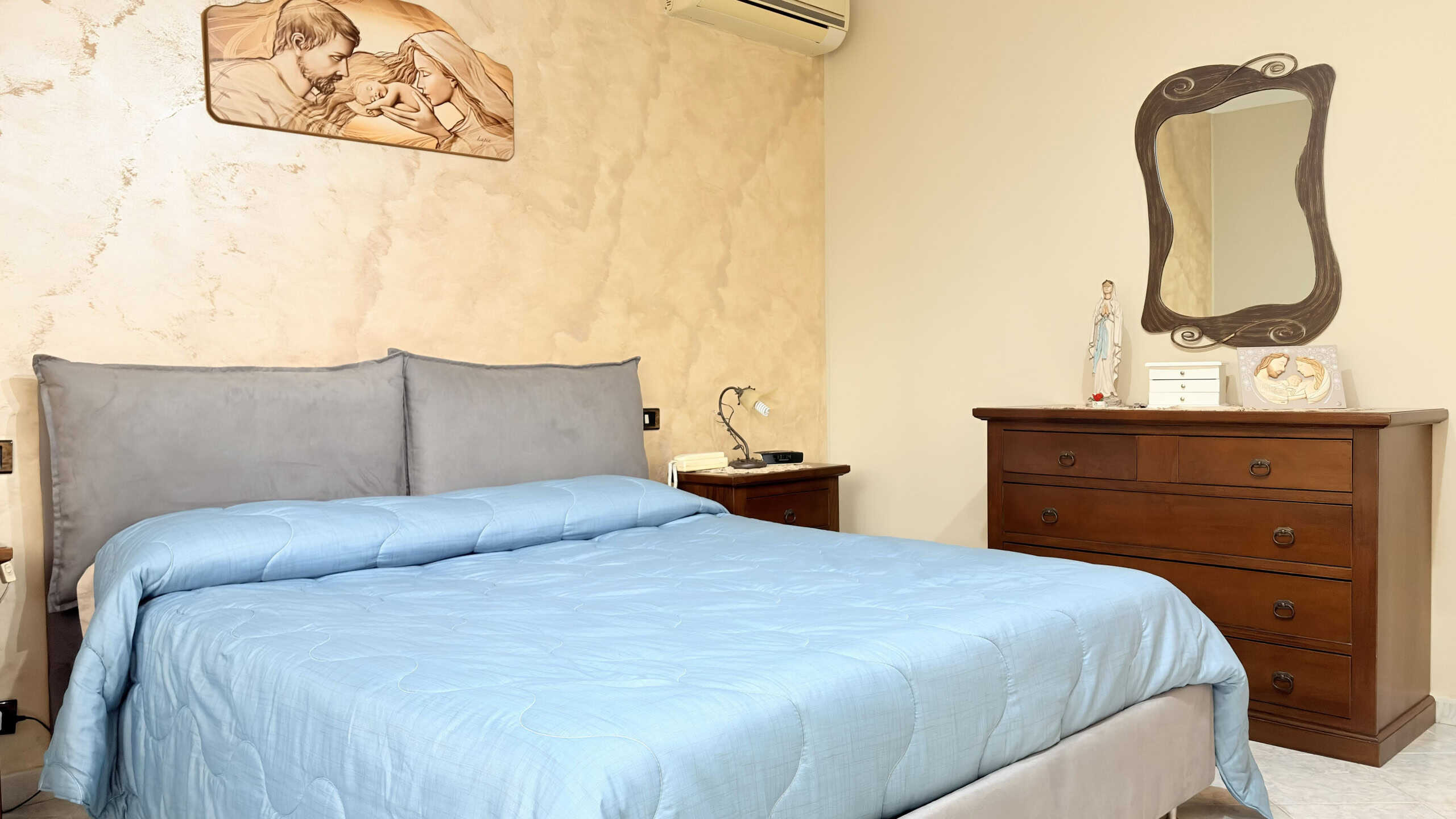 Letto matrimoniale con testiera imbottita in tessuto grigio, in una camera da letto accogliente e luminosa, con mobili in legno e decorazioni eleganti, ideale per ambienti di comfort e relax.