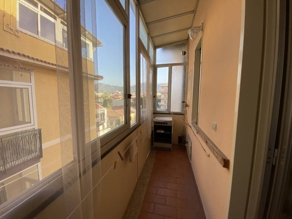 Balcone chiuso con grandi finestre, vista sulla città, ambiente luminoso e accogliente, ideale per relax o piccoli spazi verdi, situato in un edificio residenziale nel centro urbano.