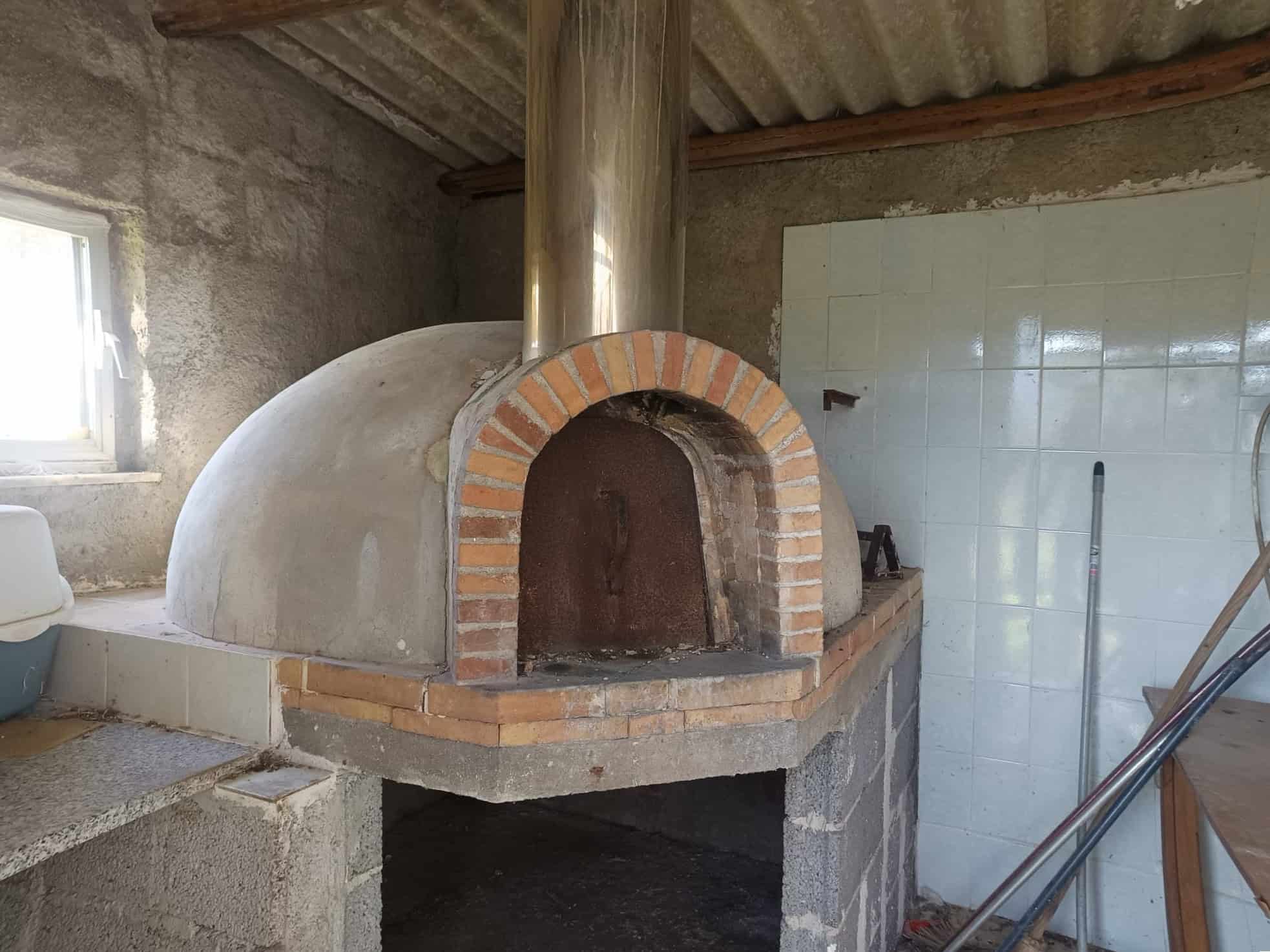 Forno a legna rustico in mattoni e cemento, ambiente di cucina con pareti in piastrelle bianche e finestra naturale, ideale per cottura tradizionale e convivialità.