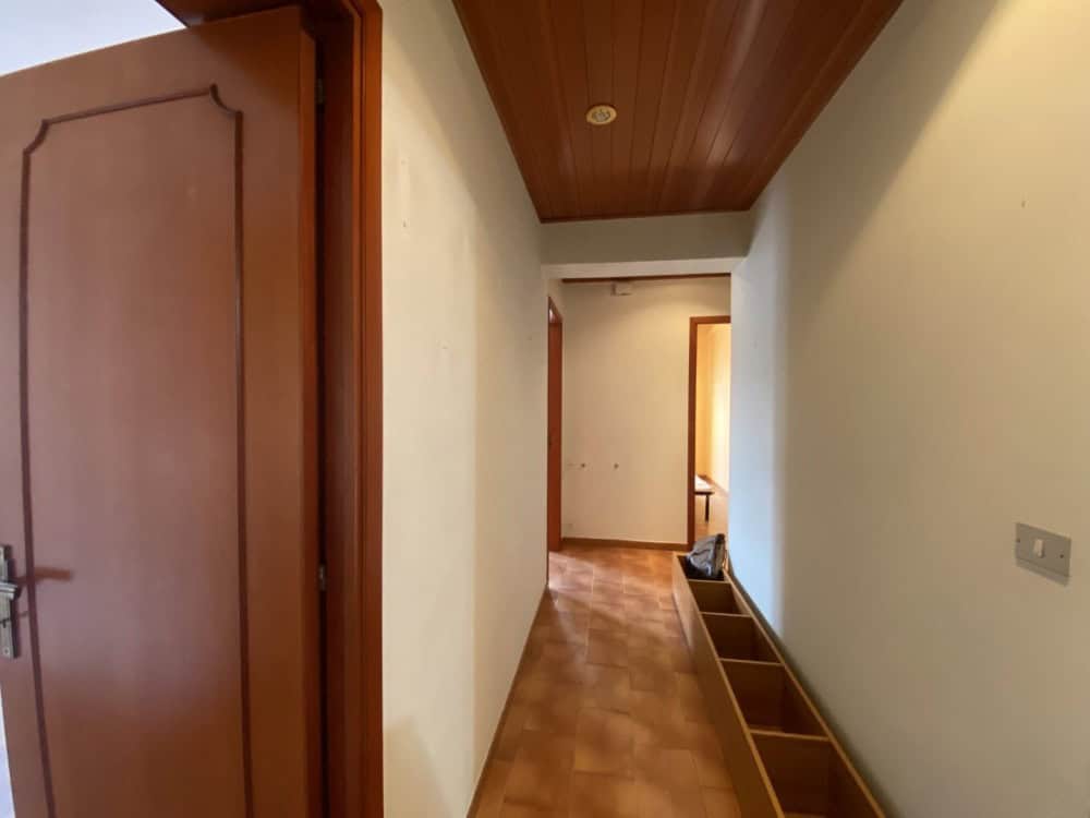 Corridoio interno di un appartamento con pareti bianche, pavimento in parquet e soffitto in legno, vista verso una stanza luminosa.