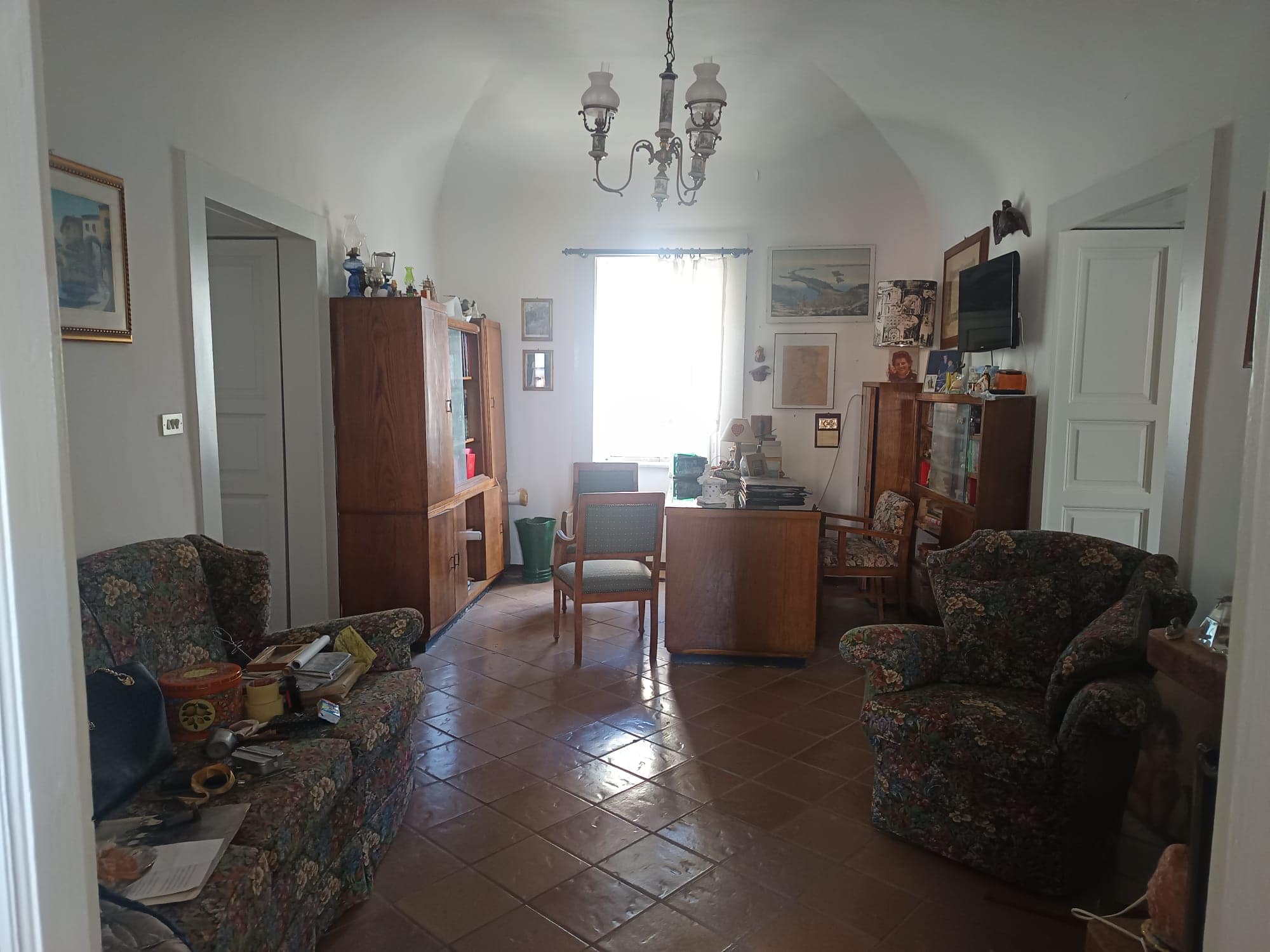 Una stanza soggiorno accogliente con arredi classici, divani e poltrone, librerie e un tavolo centrale, illuminata da una finestra con tende leggere e un lampadario antico, in stile tradizionale.