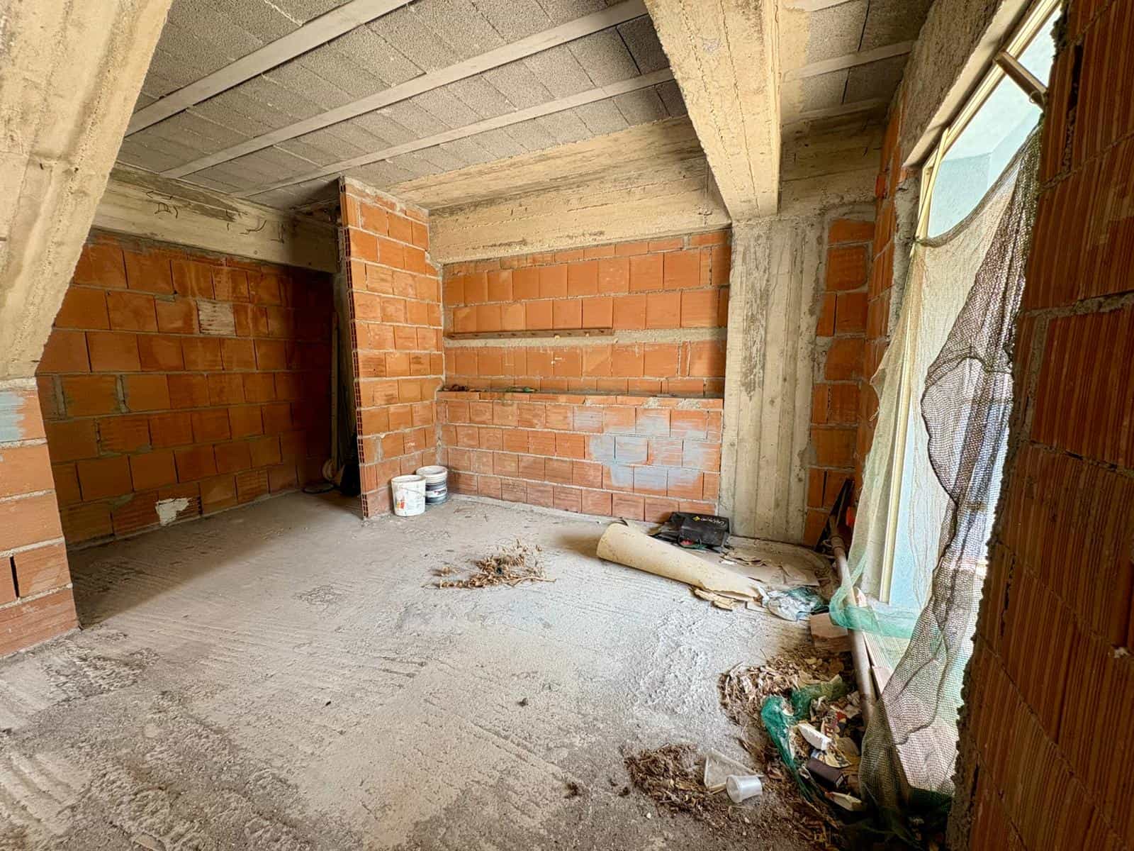 Cortile interno di un edificio in costruzione con pareti in mattoni rossi e soffitto in cemento, attrezzi e materiali di costruzione sul pavimento, luce naturale che entra da una finestra coperta da p.