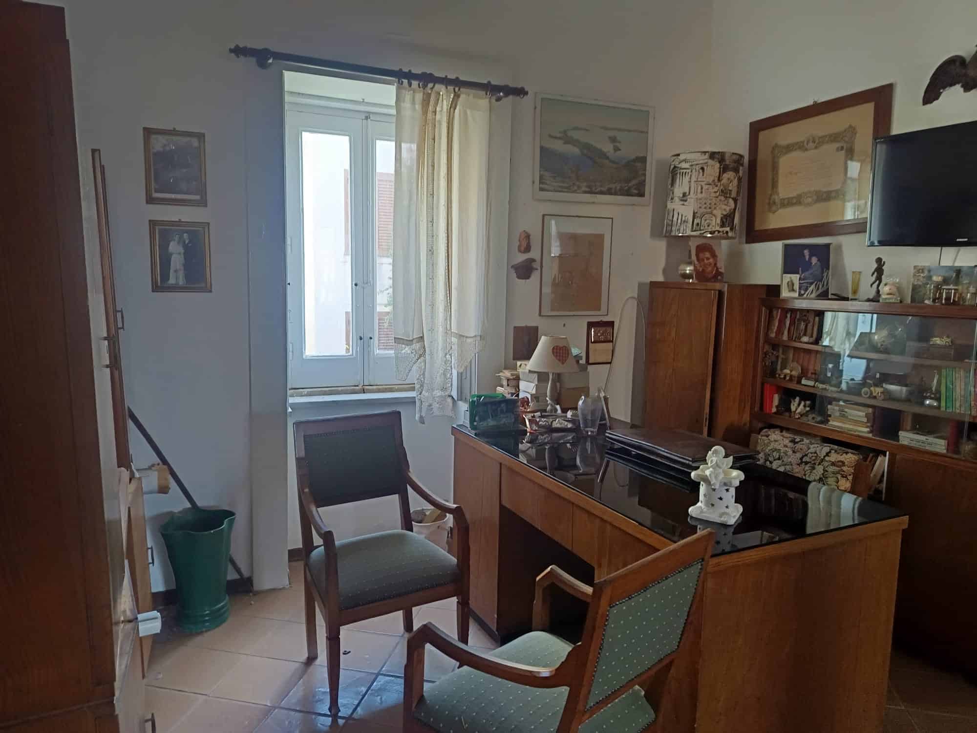 Elegante soggiorno con mobili in legno, scrivania e poltrone, decorazioni artistiche e una finestra luminosa con tende leggere, ideale per vivere in comfort e stile.