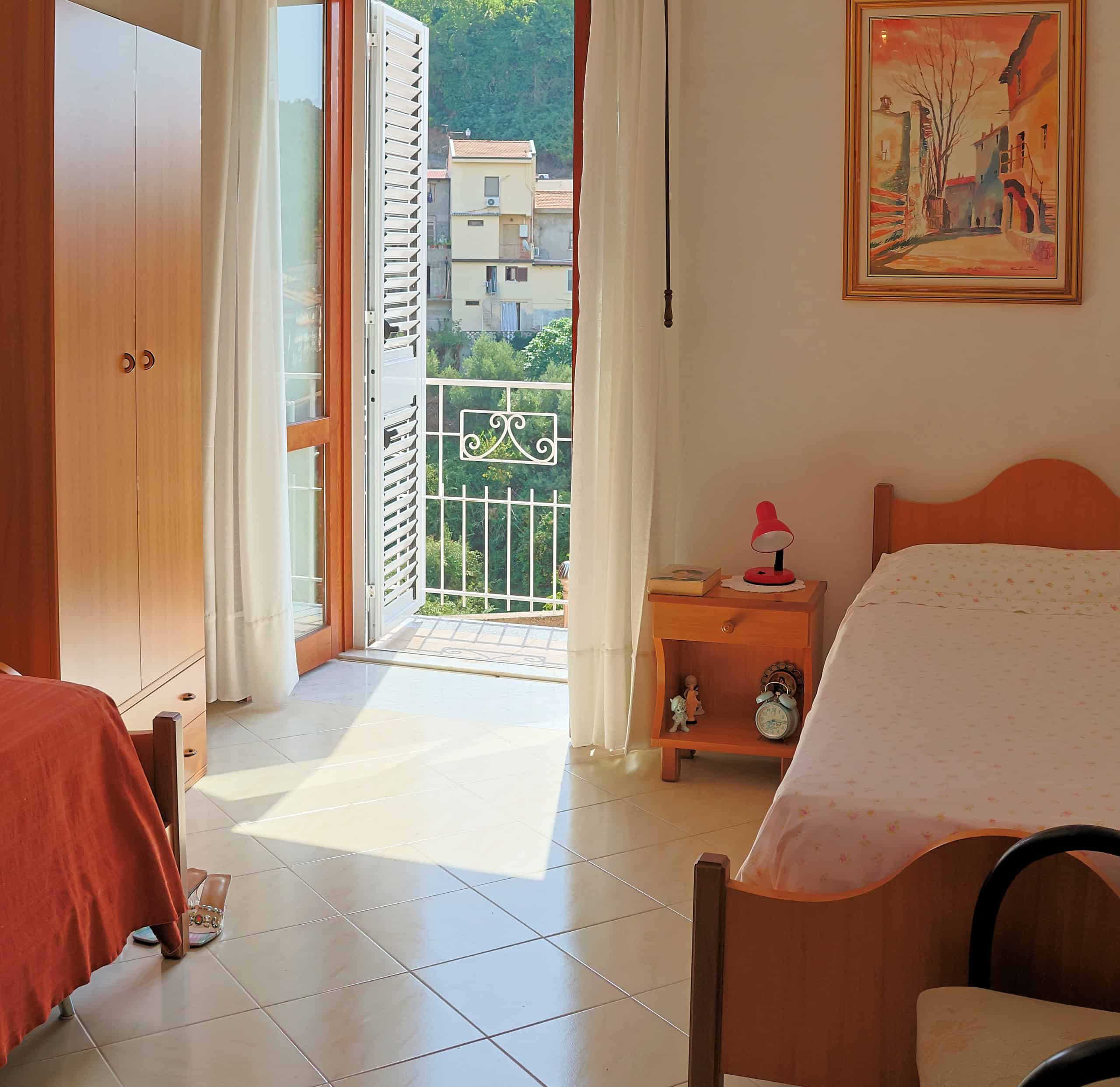 Camera da letto luminosa con balcone, arredi in legno e decorazioni colorate, vista su edifici e natura, ambiente accogliente e tranquillo.