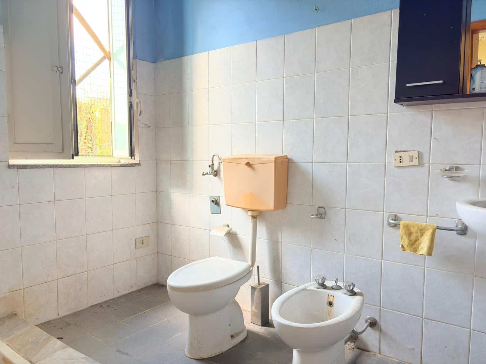 Vaschetta e bidet in un bagno semplice con pareti rivestite di piastrelle bianche e finestra aperta, illuminando lo spazio naturale e arioso.