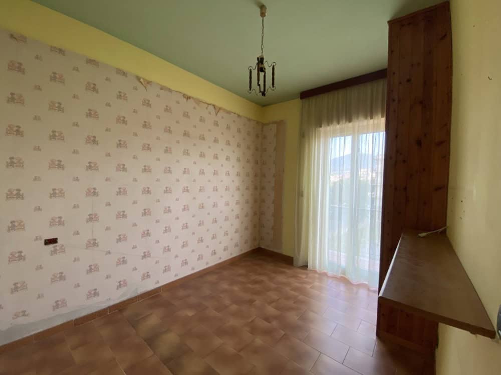 Aria luminosa e accogliente con pareti decorate, grande finestra con tende trasparenti, pavimento in piastrelle e soffitto con lampadario, ideale per soggiorno o studio.
