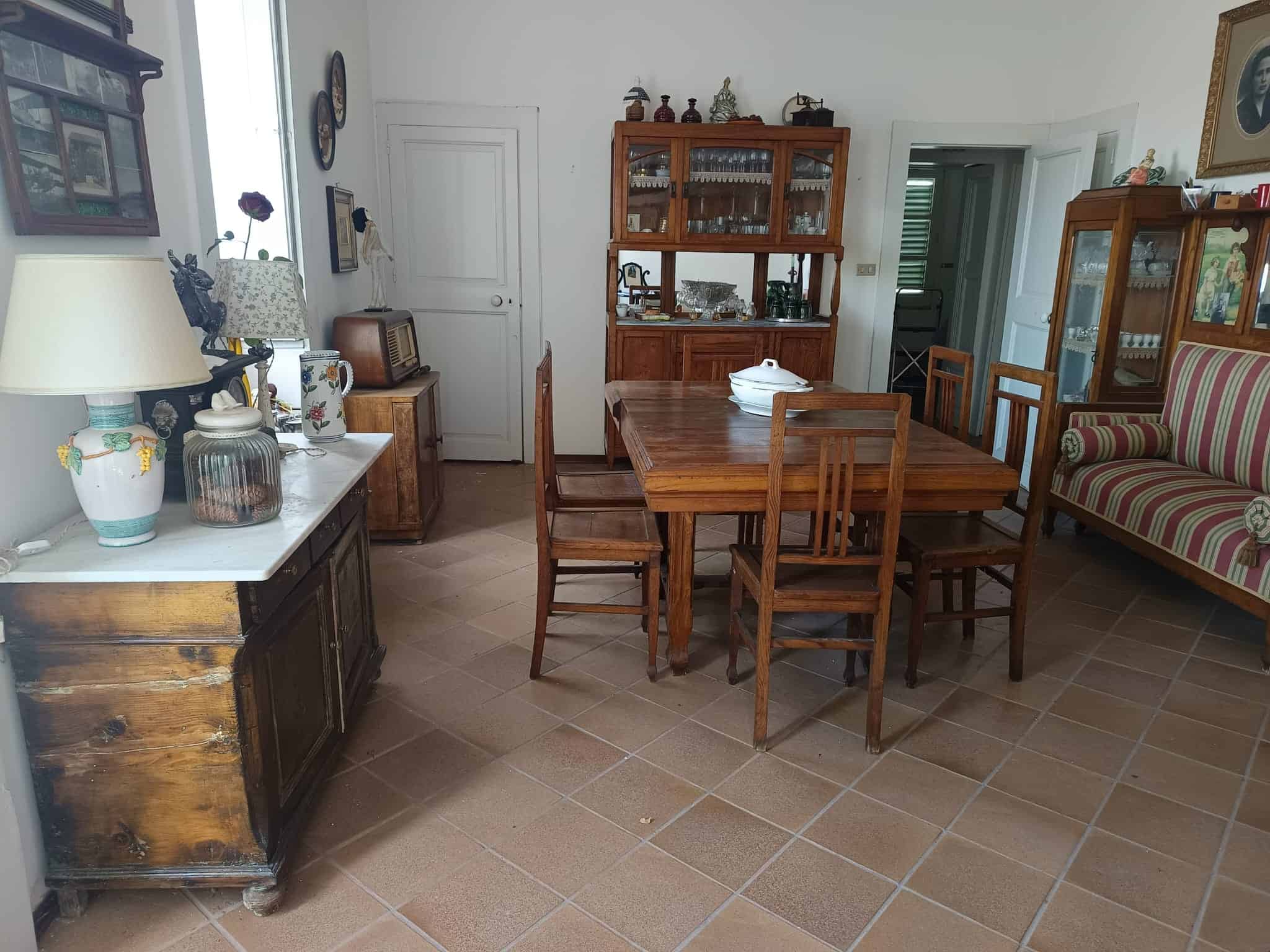 Una sala da pranzo accogliente con mobili in legno, tavolo centrale e sedie, arredamento vintage e armadi con stoviglie, illuminata da luce naturale, ideale per ambienti familiari e conviviali.
