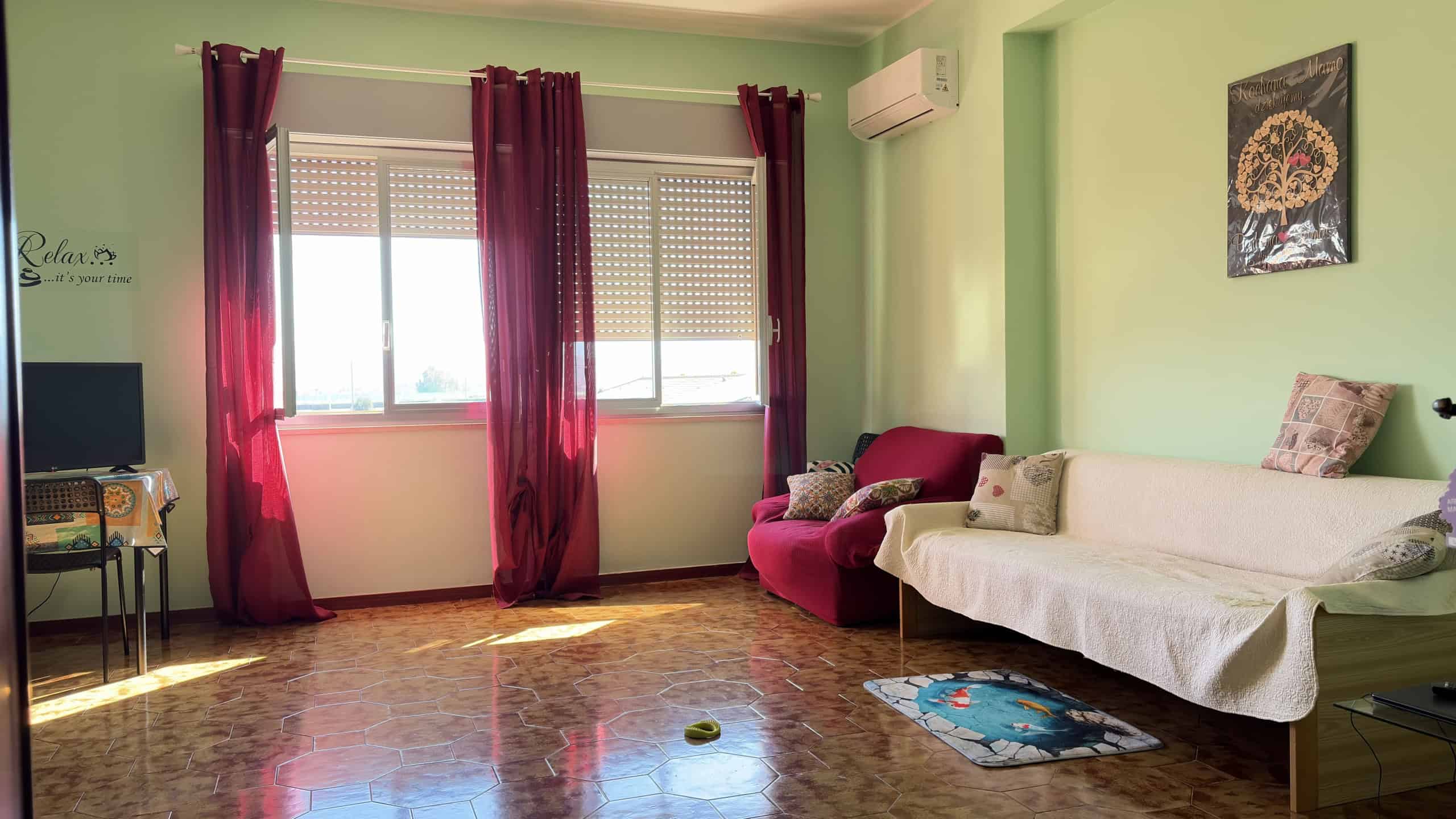 Ampia stanza soggiorno luminosa con pareti verde pastello, divano rosso e divano letto bianco, grandi finestre con tende rosse, aria condizionata e decorazioni floreali, ambiente accogliente e luminos.