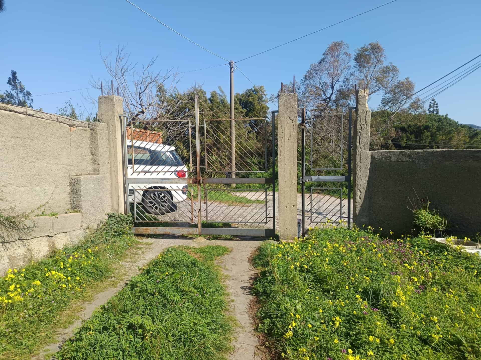 Cancello in metallo con pilastri in cemento, ingresso di un giardino con strada sterrata e vegetazione verde, ambiente esterno in zona residenziale.