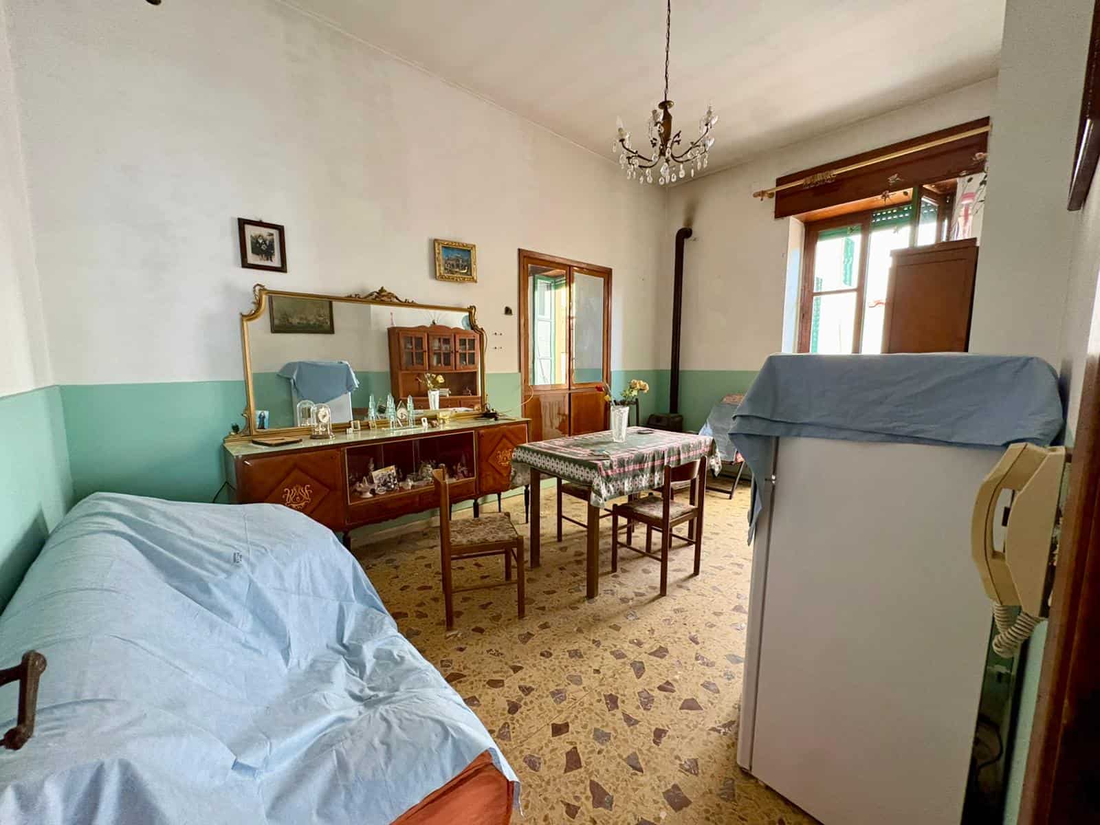 Una stanza da pranzo e soggiorno in stile classico con mobili in legno, tavolo con tovaglia, divano coperto con telo azzurro, e finestre con tende di legno, ambiente accogliente e luminoso.