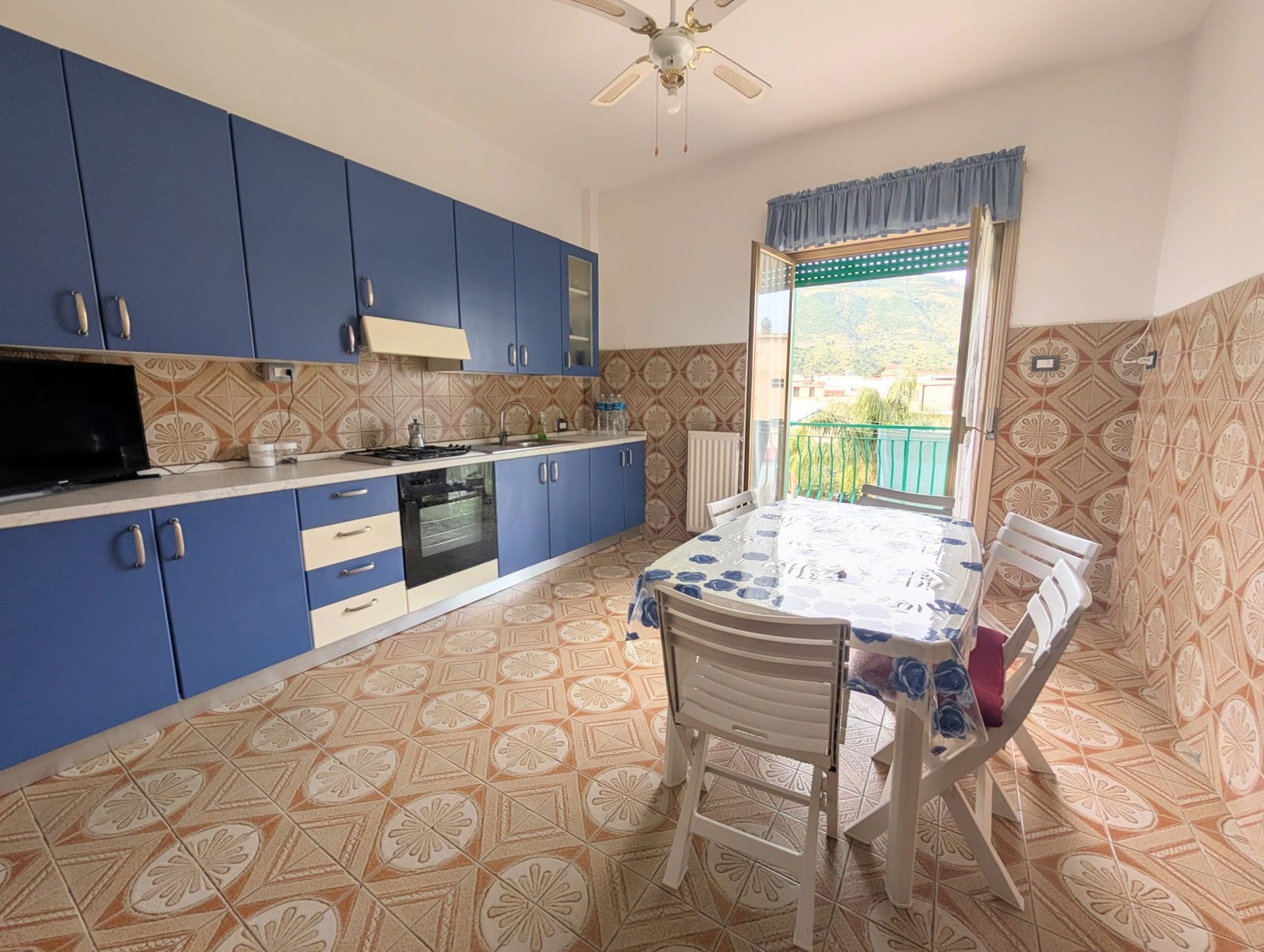 Lavastoviglie e cucina moderna in una cucina spaziosa con pareti rivestite di piastrelle colorate, tavolo da pranzo con sedie e accesso a un balcone con vista panoramica.