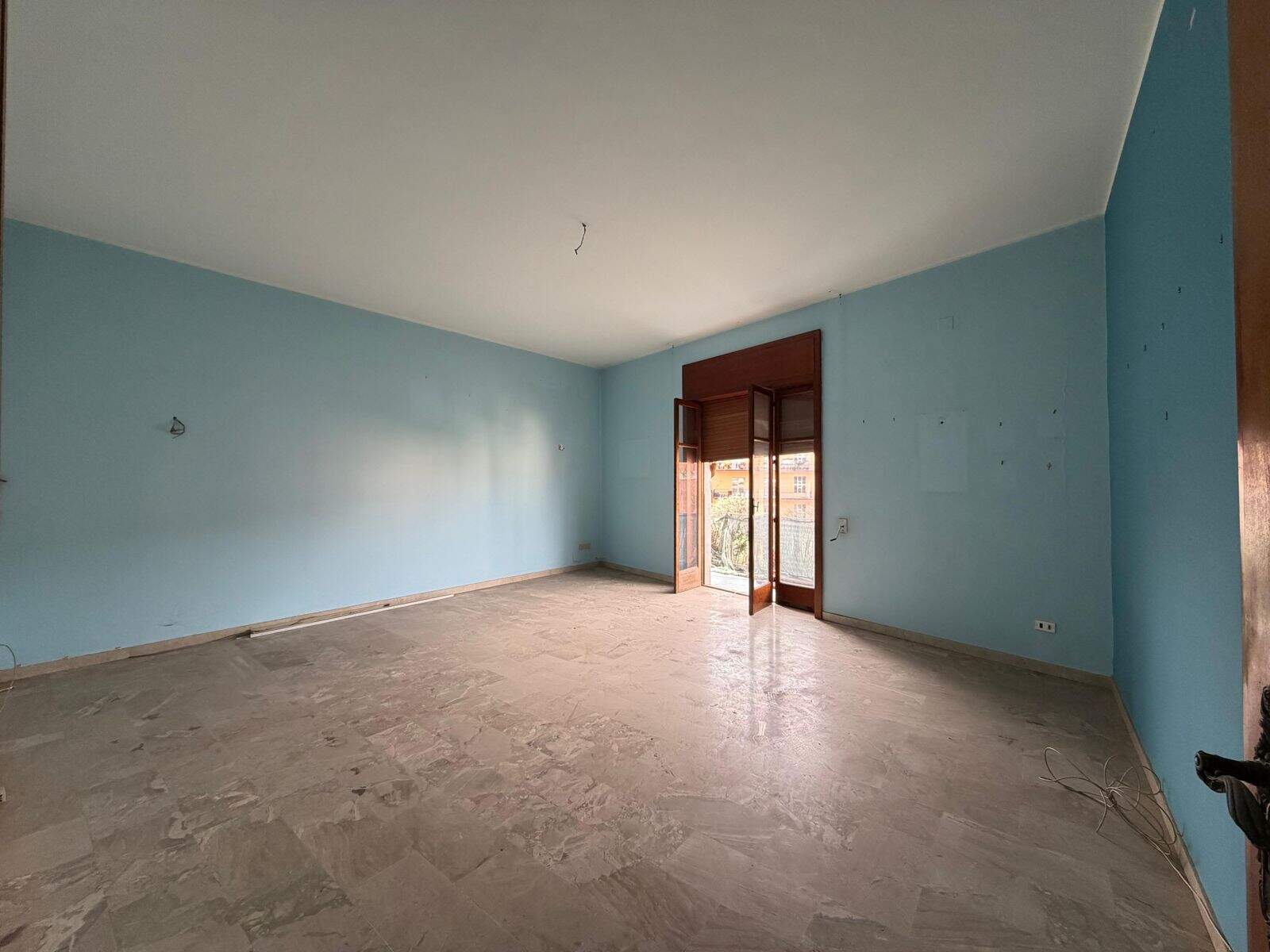 Stanza vuota in fase di ristrutturazione con pareti azzurre e porta finestra che conduce a un balcone, pronta per essere personalizzata.