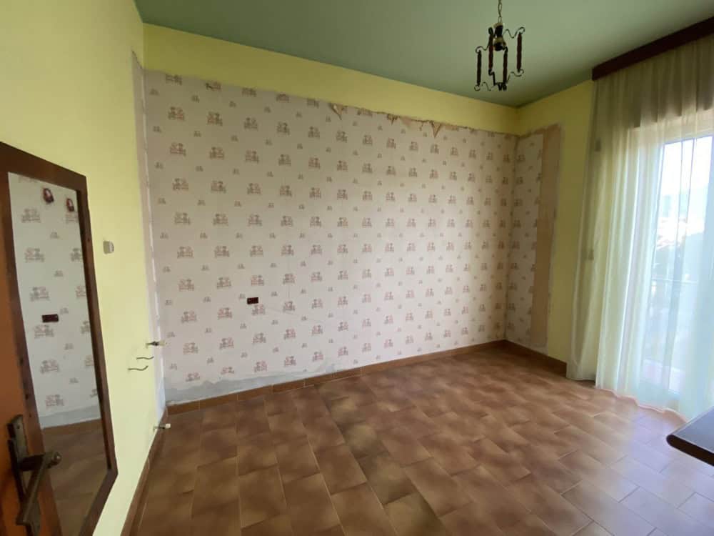 Appartamento vuoto con parete decorativa, parquet e grande finestra con tende trasparenti, luce naturale, ideale per affitto o vendita, in zona residenziale.