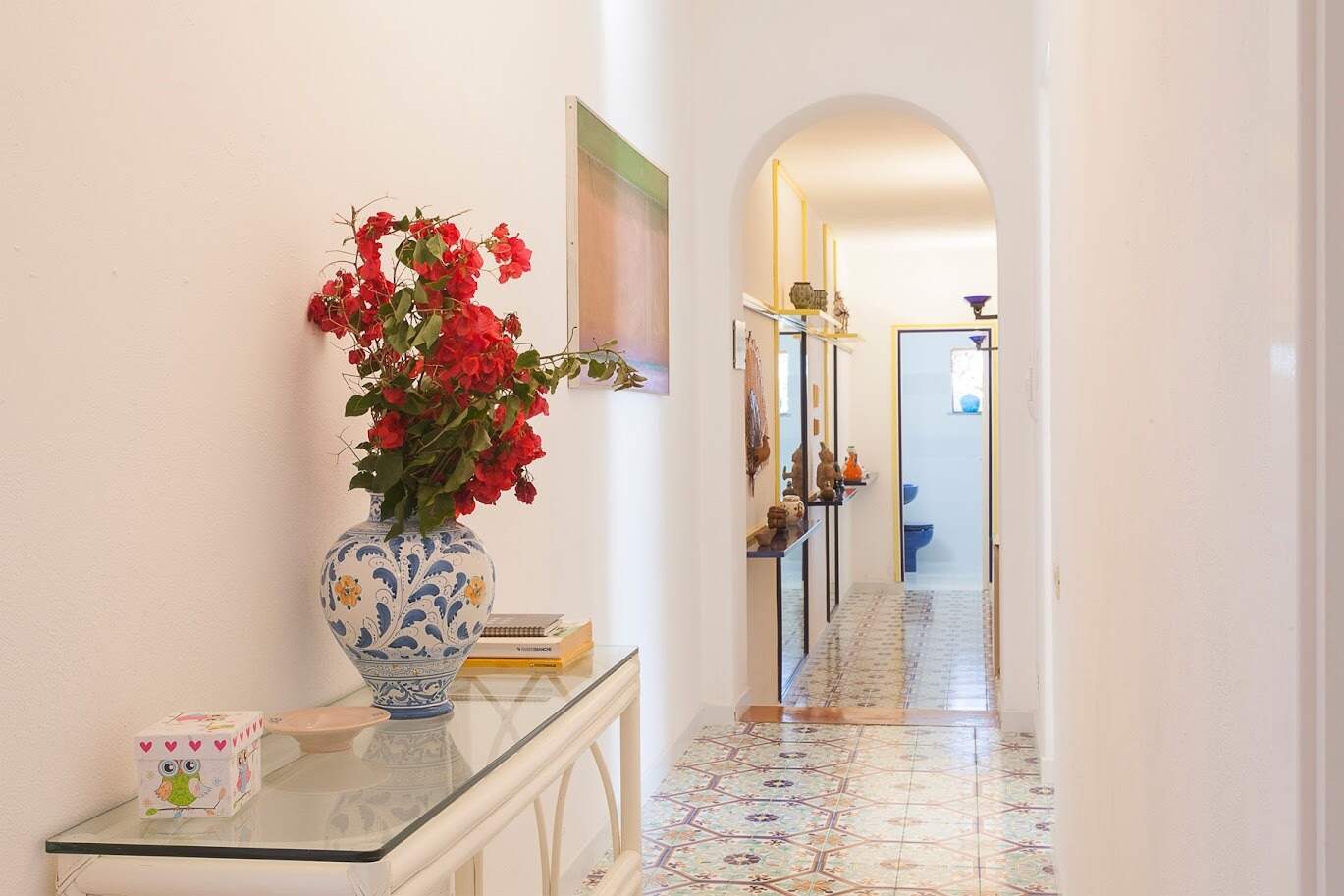 Elegante ingresso di casa con vaso di ceramica decorato e fiori rossi, corridoio luminoso con arredi raffinati e pavimento in piastrelle colorate, stile accogliente e raffinato.