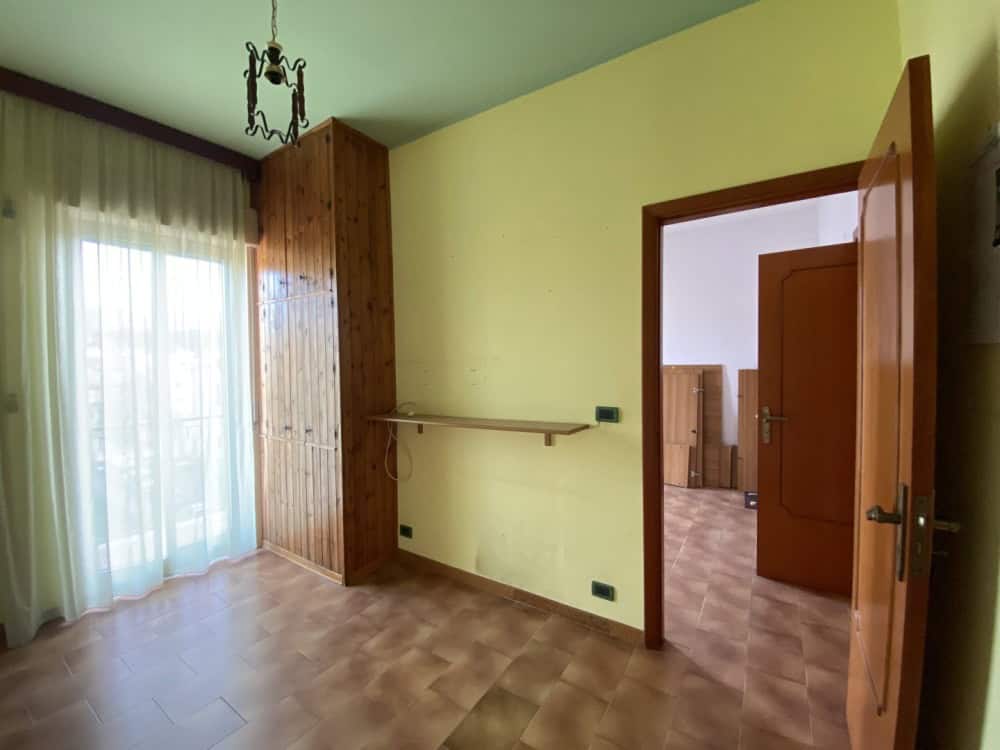 Ampia stanza luminosa con pareti verde chiaro, grande finestra con tende leggere, pavimento in parquet e arredi in legno, ideale per soggiorno o camera da letto.