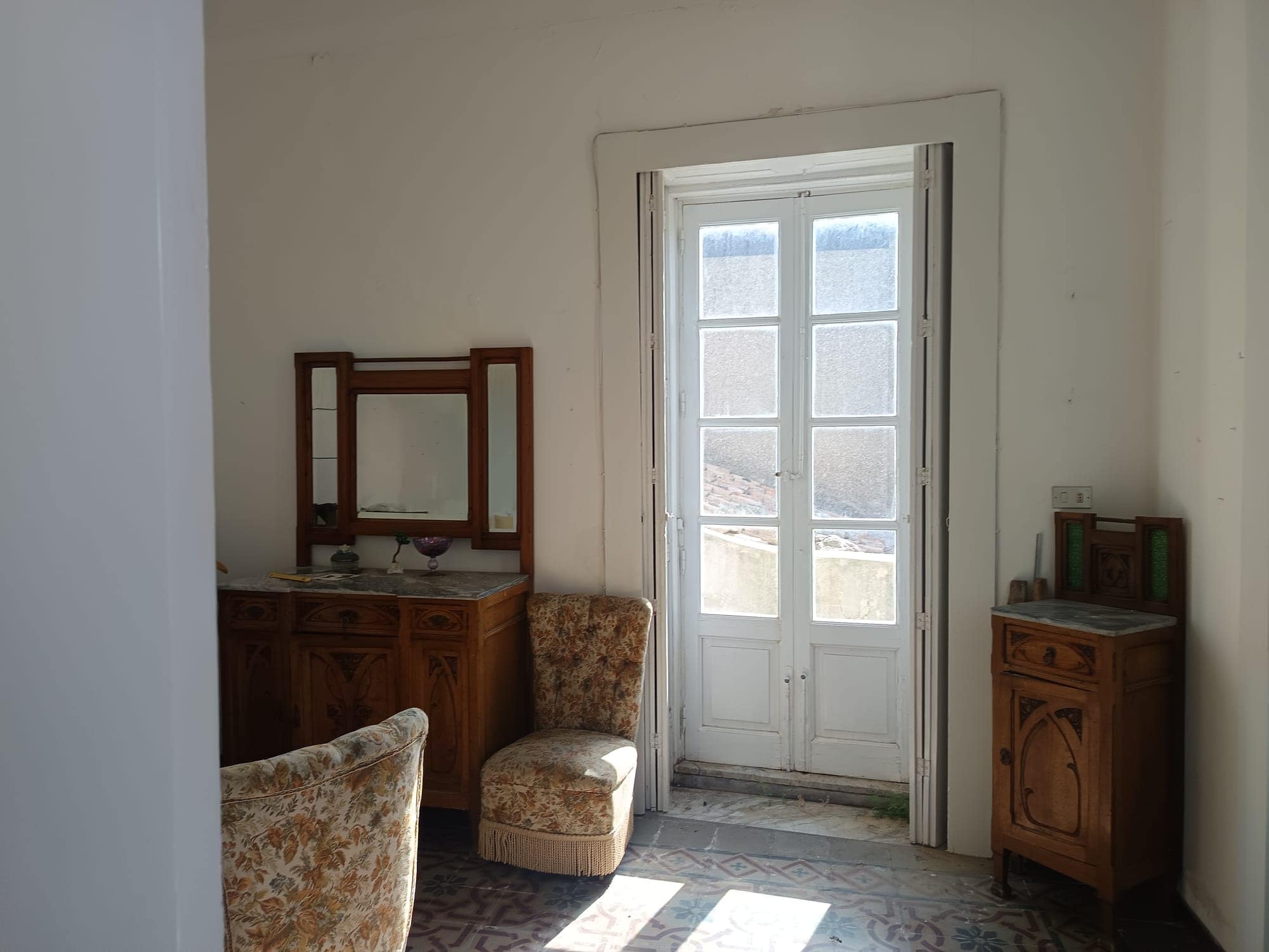 Antistante a una porta finestra in stile classico, arredamento vintage con mobili in legno e sedie imbottite, ambiente luminoso e accogliente, ideale per interni di case storiche o rustiche.